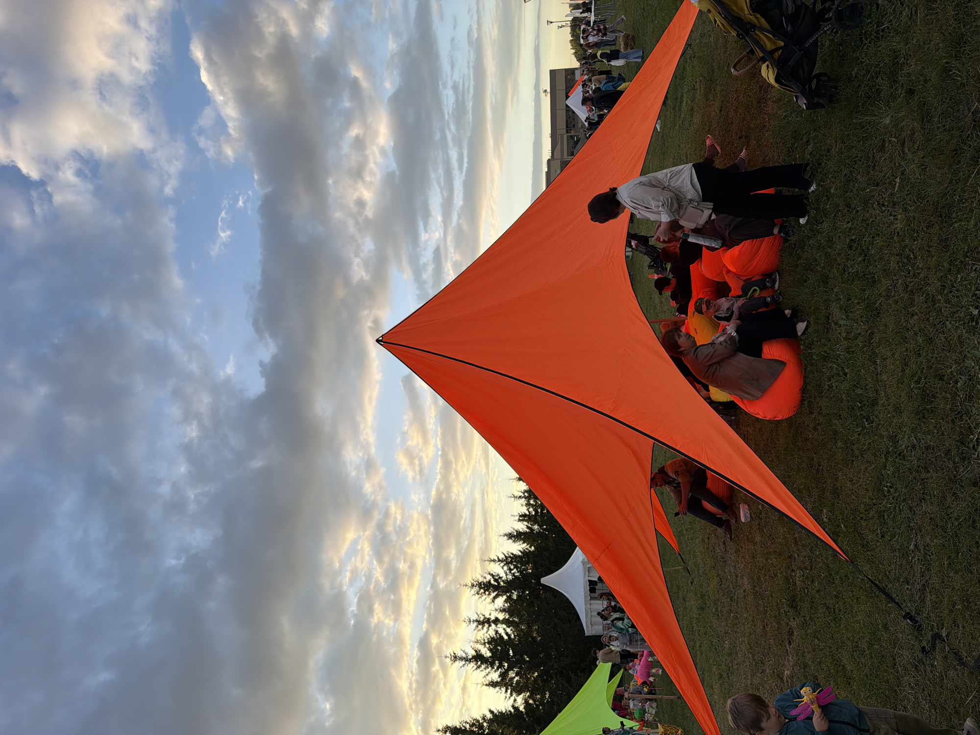 An outdoor event is in progress under a cloudy sky with patchy sunlight illuminating the scene. A large, bright orange, pyramid-shaped tent is centrally positioned, casting a shadow over a group of people seated on beanbag chairs. A woman in a light-colored jacket and black pants stands near the center of the tent, looking down at something in her hands. To the left of the orange tent, a lime green tent is partially visible, and beyond that, a white tent. In the background, a line of evergreen trees creates a dark silhouette against the sky. A few people are visible near the white tent and further in the distance, suggesting a larger gathering. A young boy in a blue jacket and khaki pants stands to the far left of the frame, holding a pink and yellow toy. A yellow stroller is parked on the right side of the orange tent. The ground is covered in green grass.