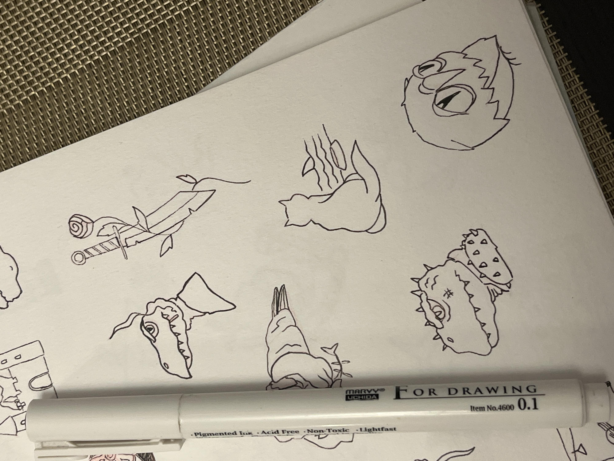The image shows a piece of white paper with several cartoon-style drawings in black ink. The drawings include an angry-looking owl, a cat curled up next to some vertical lines that may represent bamboo, a sword with a rose on the hilt and leaves on the blade, a reptilian head with a frill, a creature with a long body and rabbit-like ears covered in spines, and a creature with a spiky head and teeth. A white "Marvy Uchida For Drawing" pen lies across the bottom of the paper. The background includes a woven texture, suggesting the paper is on a table or surface with a similar pattern.