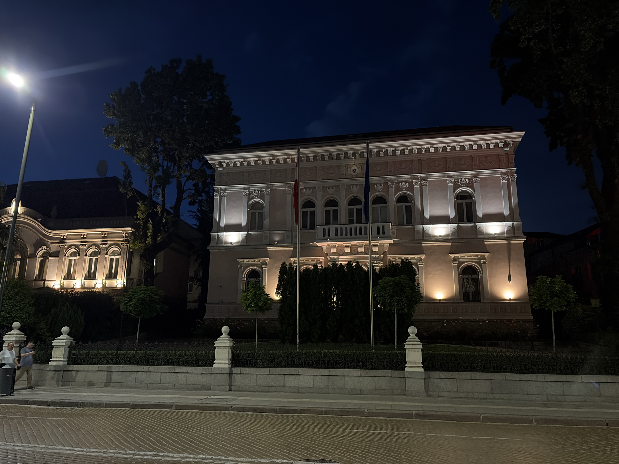 This nighttime image depicts the Presidential Palace in Sofia, Bulgaria. The central two-story building features a light-colored, ornate facade with classical architectural elements, including decorative friezes, cornices, pilasters, and arched windows. The building is illuminated by warm, upward-facing spotlights. A central balcony with a balustrade is visible on the second floor. Two flagpoles stand in front; one displays the blue flag of the European Union with yellow stars, and the other a flag predominantly red with a narrow vertical white stripe near the hoist. In the foreground, a light-colored cobblestone street borders a sidewalk. A low stone wall with decorative pillars and a metal fence separates the sidewalk from manicured bushes and small topiary trees. Two men are present on the far left of the sidewalk: one stands near a dark trash receptacle, and the other walks rightward, looking at a mobile phone. A bright, cool-toned street lamp illuminates the far left, partially revealing another building with similar architecture. Large, dark trees silhouette against the dark blue night sky, which shows faint cloud patterns.