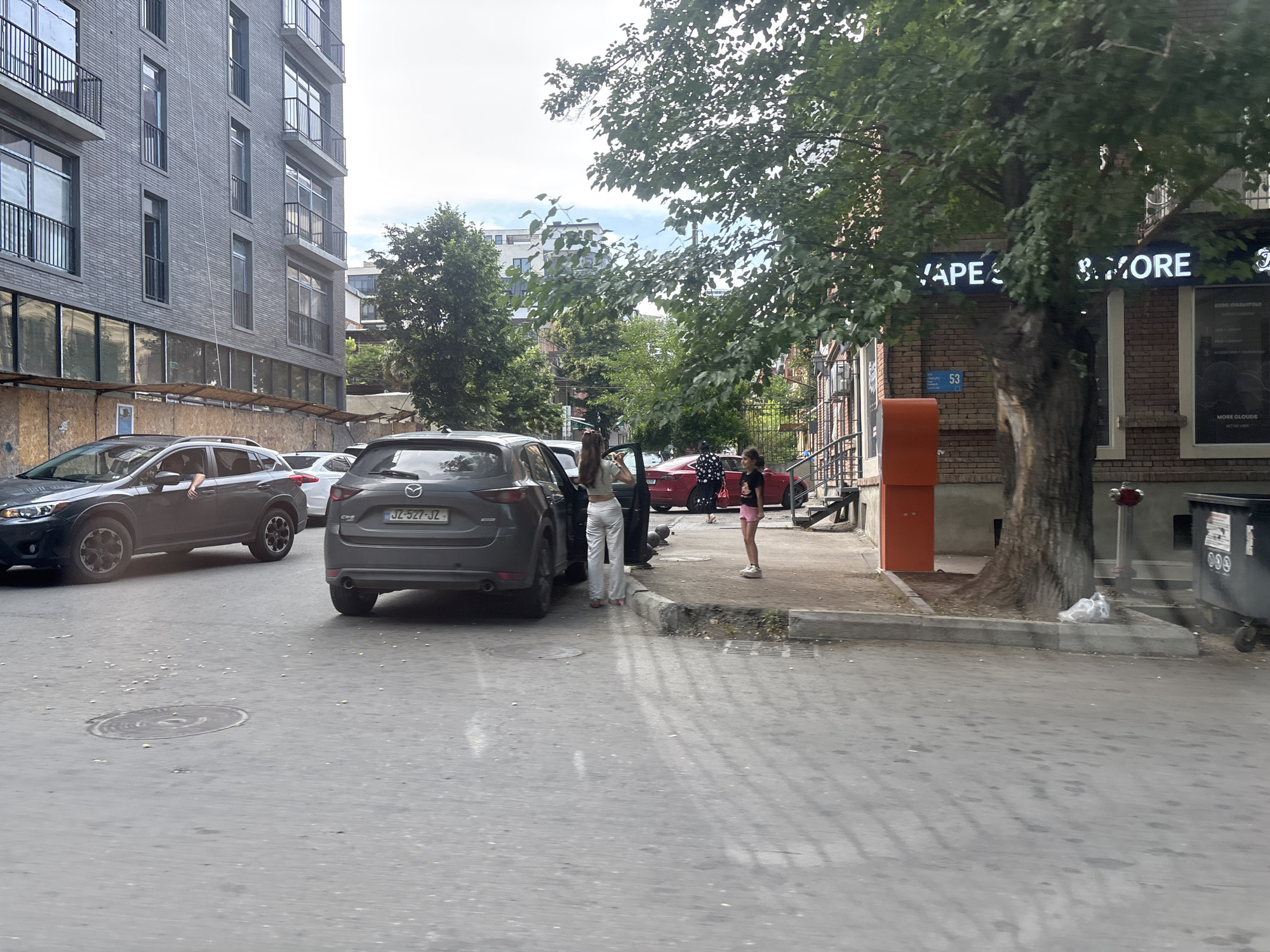 A street scene in Tbilisi, Georgia, features a gray SUV parked on the right side of the frame with its rear facing the viewer. A dark gray Mazda CX-5 is parked to its left, with its driver's side door open. A person wearing white pants and a light-colored top stands by the open door, facing away from the camera. To the right of the Mazda, a red car is parked. A young girl in a pink skirt and black top stands on the sidewalk, looking towards the cars.
The background shows a multi-story brick building with multiple windows and balconies, and a tree with dense foliage partially obscuring the building. A street sign reading "53" is visible on the brick building. Another building with large windows and signage displaying "VAPE & MORE" is also present. A large black dumpster is situated on the right side of the frame. The ground is paved with asphalt. The overall impression is a sunny day, given the shadows. The scene appears to be a typical urban street.