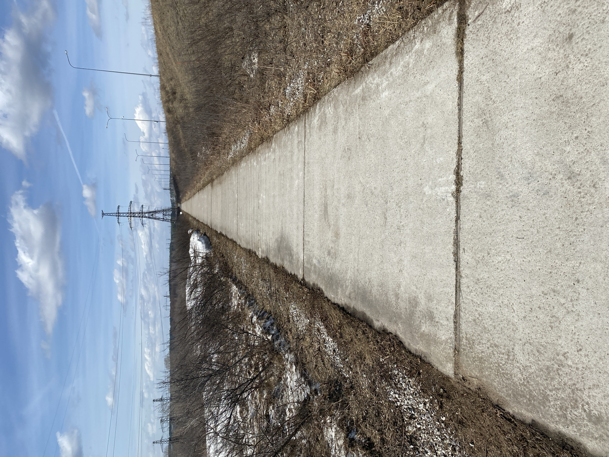 The image depicts a concrete pathway stretching into the distance. On the right of the path is dry vegetation and gravel. On the left, the view includes power lines, utility poles and a blue sky with clouds. Patches of snow are visible alongside the vegetation. The location is the city of Chaikovsky, Russia.