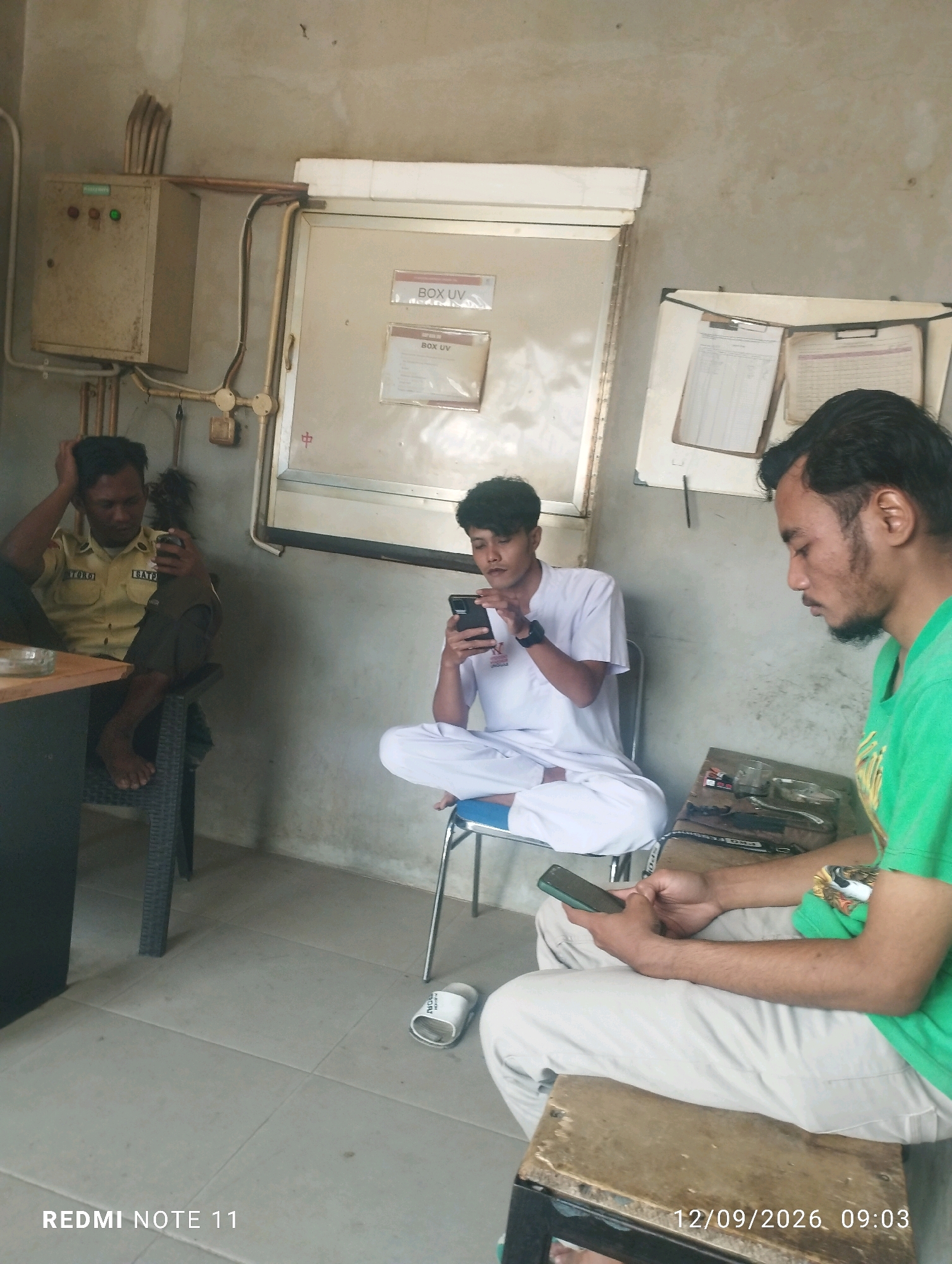 The image depicts an indoor scene with three individuals. In the foreground on the right, a man wearing a green t-shirt and light-colored pants sits on a stool. He is holding a smartphone in his right hand and appears to be looking at it. To his left and slightly further back, a man in a white outfit, possibly a uniform or religious attire, is seated on a blue-metal chair. He is also engrossed in his smartphone, holding it with both hands. On the far left, a man wearing a tan uniform with patches on the sleeves and dark pants sits on a dark wicker chair. He is looking at his smartphone, which is held in his right hand, while his left hand is raised to his head, as if in thought or discomfort.
The environment appears to be a utilitarian space. A grey concrete wall serves as the backdrop. To the left, a cream-colored electrical control panel with several lights is mounted on the wall. To the right of this panel, a large, beige cabinet with two signs indicating "BOX UV" is visible. A white whiteboard with papers attached is mounted on the wall above and to the right of the cabinet. The floor is tiled with light gray tiles. There is a wooden table on the left side of the image, and a small stack of items is on the table near the seated man in uniform. A white slipper with the word "BOK" is visible on the floor in the center right of the image. The image also has overlaid text in the bottom left corner reading "REDMI NOTE 11" and in the bottom right corner reading "12/09/2026 09:03".