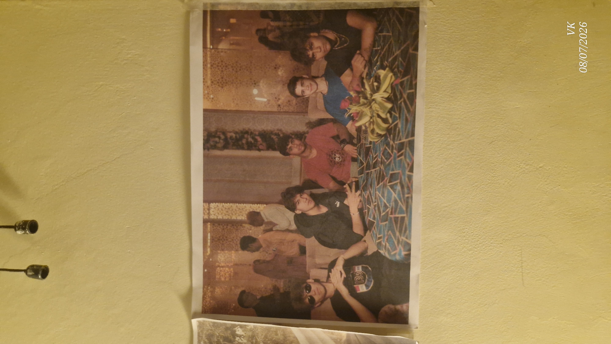 The image shows a framed photograph mounted on a textured, pale yellow wall. The photograph itself is a snapshot of a group of five young men seated around a table. The men are casually dressed. The table is covered with a patterned tablecloth, and a floral arrangement is centrally placed on it. In the background of the photograph, other people are visible, some of whom appear to be engaged in conversation. The lighting within the photograph suggests an indoor setting, possibly a restaurant or a gathering space. The wall in the foreground is bare except for a timestamp in the lower right corner that reads "VK 08/07/2026". Two dark, metallic objects, resembling light fixtures or decorative elements, are partially visible at the very top of the image, hanging from the ceiling. The overall impression is of a casual social gathering captured in a photograph.