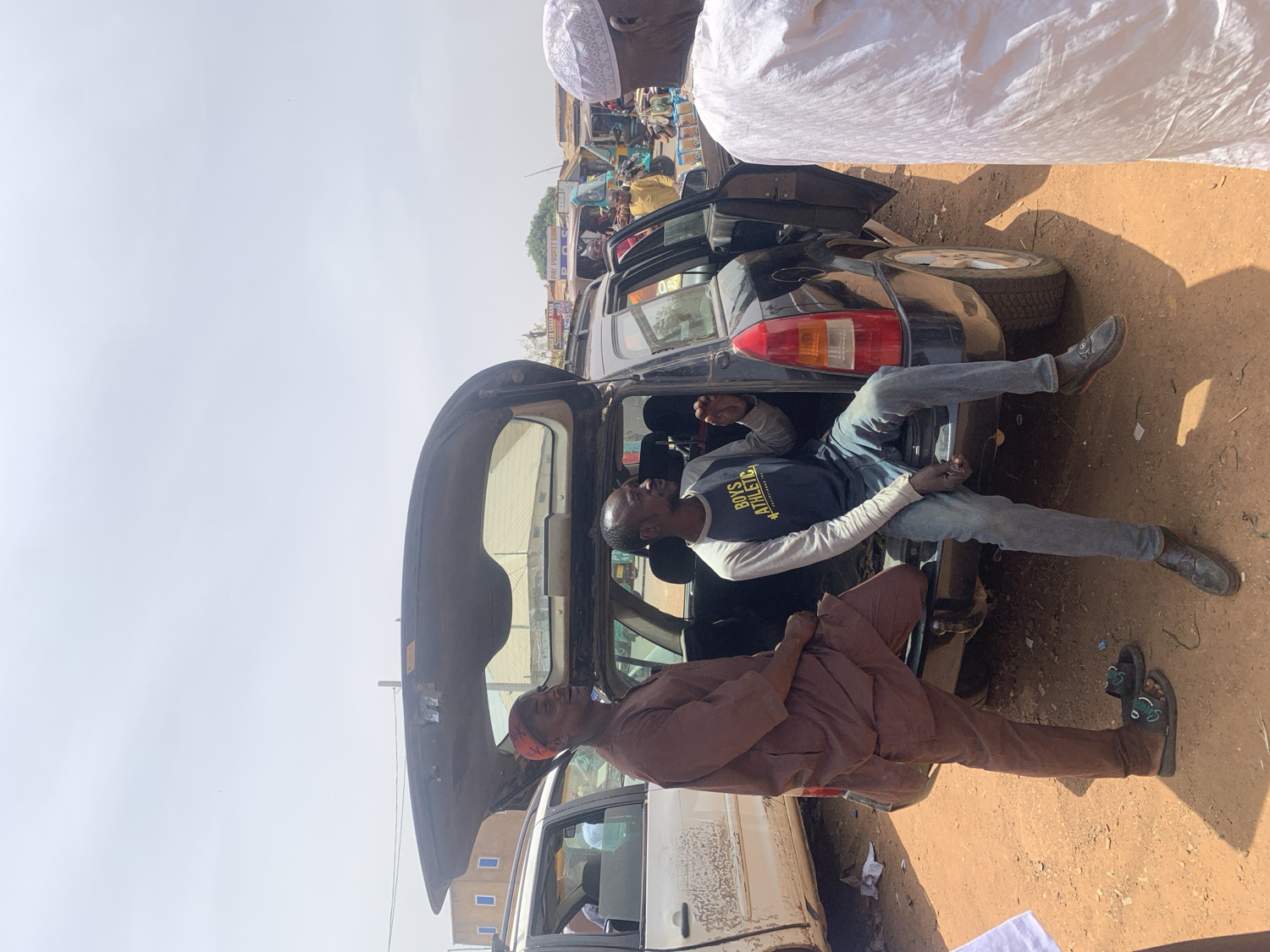 This image captures a daytime scene in Rafin Par, Nigeria, set on an unpaved, reddish-brown dirt street that appears to be a market or busy commercial area. The central focus is a dark-colored hatchback car with its rear hatch open.
Inside the open trunk, a man is seen in a reclined or resting position, with his legs dangling out. He wears a dark blue long-sleeved shirt with the text "BOYS ATHLETIC" in yellow across the chest, light blue jeans, and dark shoes. His eyes are closed or cast downwards, suggesting he might be sleeping, resting, or unconscious.
Standing next to the car, a man dressed in a brown traditional outfit, possibly an agbada, with a red cap and dark sandals, is observing the scene. In the upper right corner, another individual wearing white traditional attire and a white skullcap (kufi) is partially visible. A portion of a white car can also be seen in the bottom left of the frame.
The background reveals market stalls and structures, indicative of a bustling environment. The sky is bright and clear, signifying a sunny day. The image, rotated 90 degrees clockwise, presents a candid moment of daily life and interaction amidst people and vehicles in an active street setting.