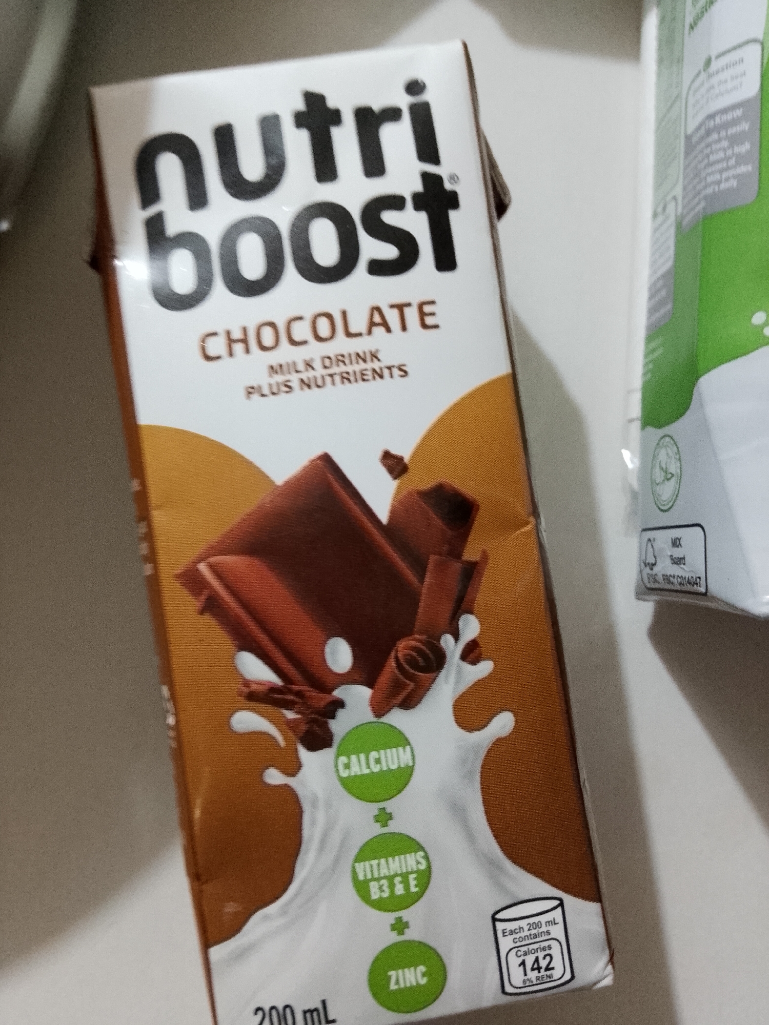 The image displays a carton of "Nutriboost Chocolate Milk Drink Plus Nutrients" in the foreground, standing upright. The carton is primarily white, featuring the brand name "nutriboost" in large black letters. Below this, "CHOCOLATE" is prominently displayed, followed by "MILK DRINK PLUS NUTRIENTS". The design on the lower part of the carton shows chocolate pieces and shavings falling into a splash of milk. Three green circular icons highlight the added nutrients: "CALCIUM", "VITAMINS B3 & E", and "ZINC". A nutritional label indicates that "Each 200 mL contains Calories 142" and "6% RENI". The carton's volume is specified as "200 mL".
To the right, a second carton is partially visible, featuring a green label that includes a "halal" symbol. Text on this carton also indicates "MIX Board" and "FSC® C014047", signifying sustainably sourced packaging. The background is light and indistinct, suggesting an indoor setting such as a shelf or countertop. No people are present, and no particular activity is taking place. The general location context for this media file is Davao City, Philippines.