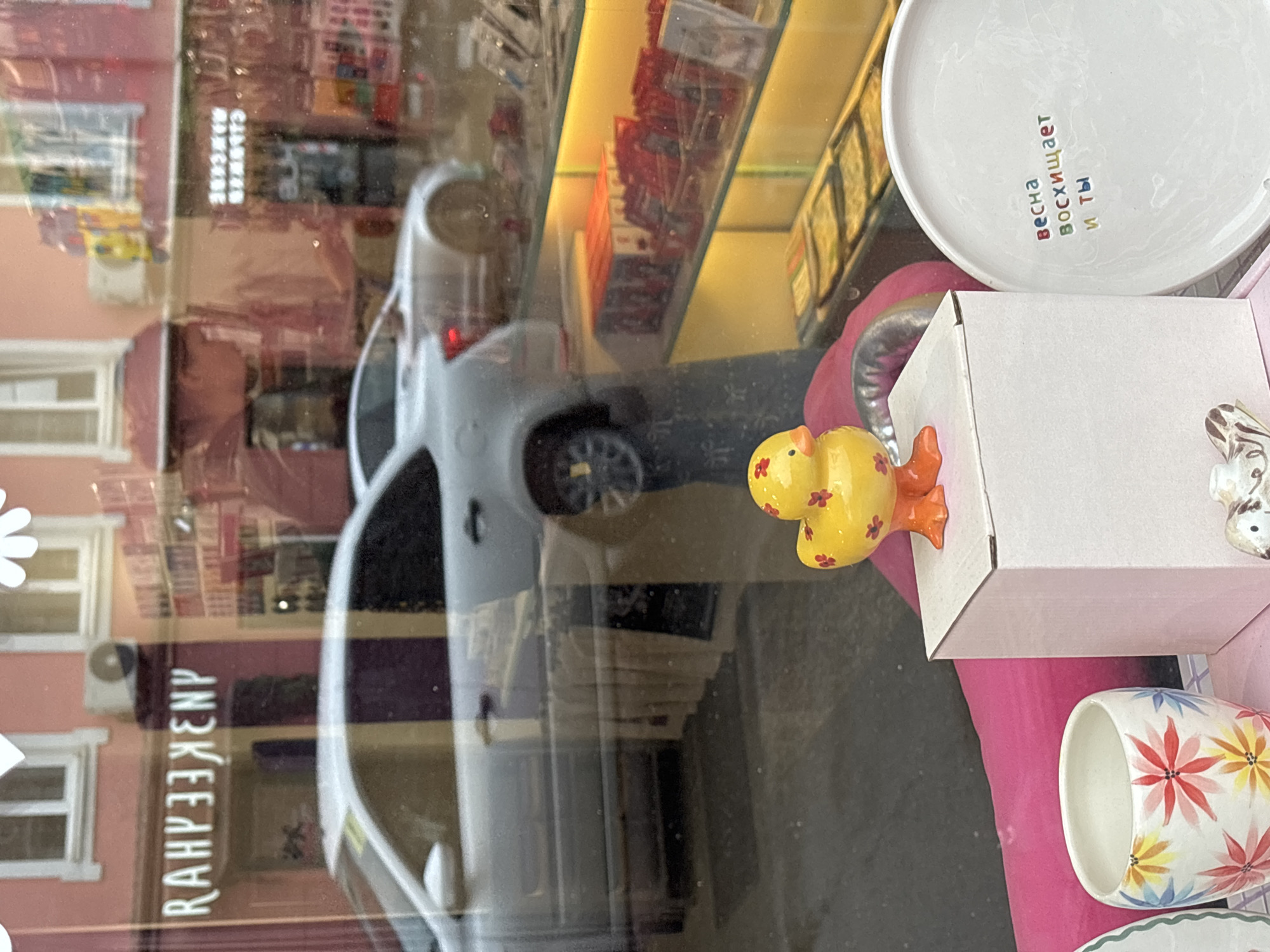 The image shows a street-level view of a shop window. A small, yellow ceramic duck figurine with red floral patterns and orange feet is displayed on a white box. To the right of the duck, a white plate is visible with colorful text in Russian, reading "весна восхищает и ты" (spring is amazing and you). A white ceramic cup with floral designs is positioned to the left of the duck. Reflected in the window are a silver car driving past, buildings with windows, and what appears to be an air conditioning unit on the facade of a pink building. The text on the building above the duck in the reflection reads "RAHYBEKENP". The overall scene suggests a retail environment, possibly a gift shop or home decor store, with merchandise displayed both inside and reflected from the outside. The location appears to be Nizhny Novgorod, Russia, based on the presence of Russian text and architectural style often found in Russian cities.