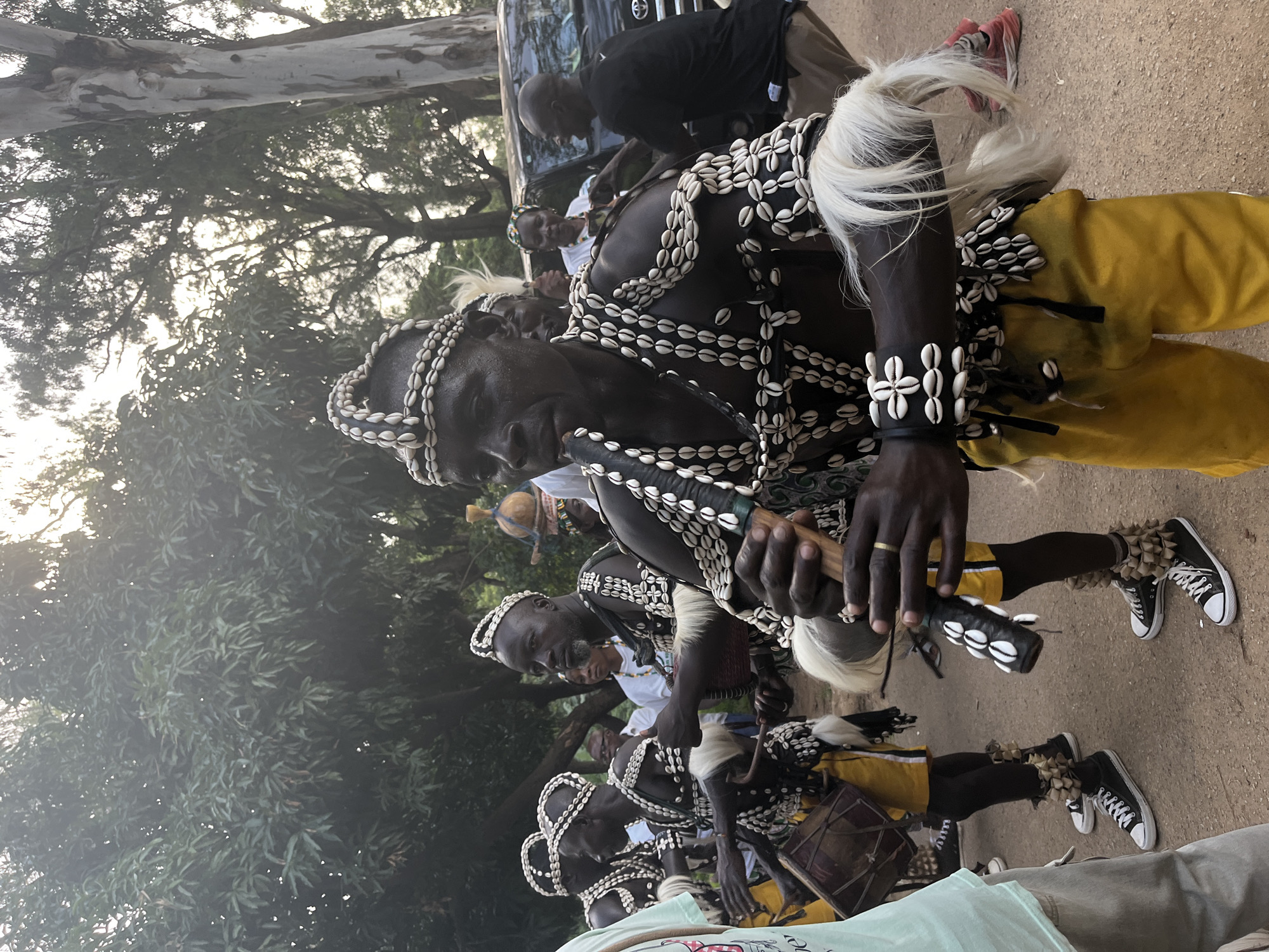 The image, taken in Jos, Nigeria, depicts a group of men dressed in traditional attire. Their outfits are adorned with numerous cowrie shells, forming intricate patterns on their chests and headdresses. Some individuals wear white fur or hair accents, adding to their ceremonial appearance. They are also wearing yellow pants and black sneakers. Several men are holding what appear to be ceremonial sticks or instruments, and some are accompanied by drums.
The scene suggests a cultural performance or ritual taking place outdoors. The lighting indicates it is either early morning or late afternoon, with soft, diffused light. The background shows trees and a dirt or gravel path, typical of an outdoor gathering space in Nigeria. The expressions on the faces of the men are serious and focused, suggesting the importance of the event. The overall atmosphere is one of tradition and community engagement. No visible text is present in the image.