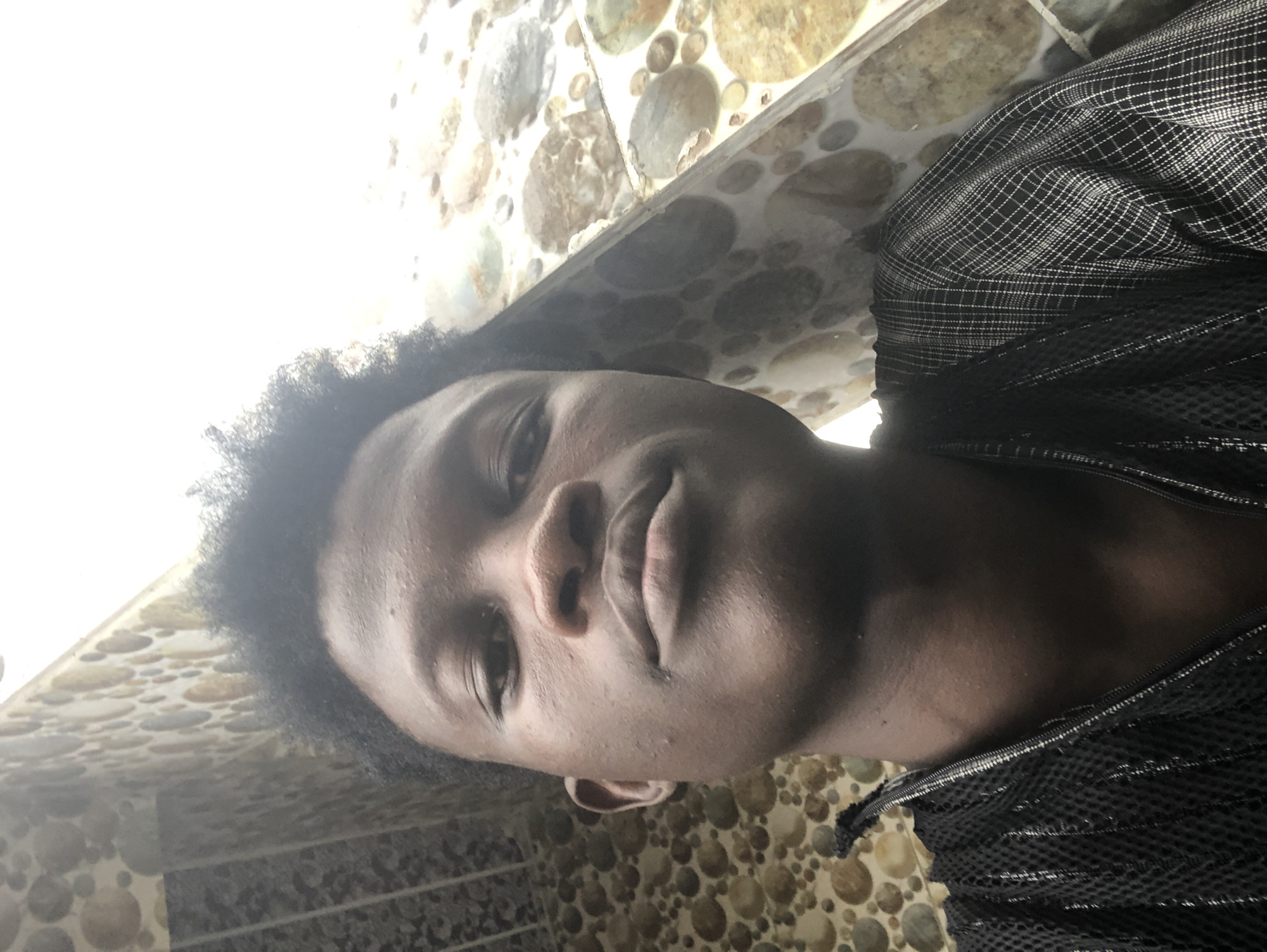 A young Black male is taking a selfie. He is looking directly at the camera, with his head tilted slightly upwards. The background consists of a wall with a decorative pattern of circular, stone-like tiles. He is wearing a black garment with a textured, mesh-like material on the left side and a checkered pattern on the right side. The lighting suggests the photo was taken outdoors during the day, with bright light coming from the upper right of the frame. The scene appears to be in a residential area in Ado, Nigeria, given the style of the tiled wall.