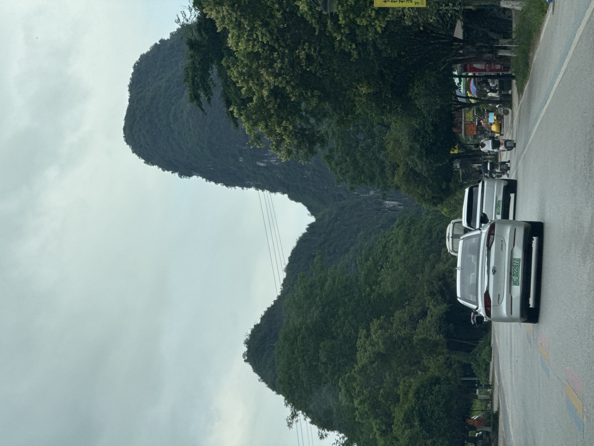 The scene depicts a road in a likely rural area near Guilin, China. The road has two silver cars, oriented in the same direction of travel. There is a small grouping of mopeds and people along the right side of the road further up towards the top of the photo. A prominent mountain forms a backdrop, with lush trees lining the road and base of the mountain. Overhead power lines are visible. The sky is overcast. The license plate on the car is 桂C022877.