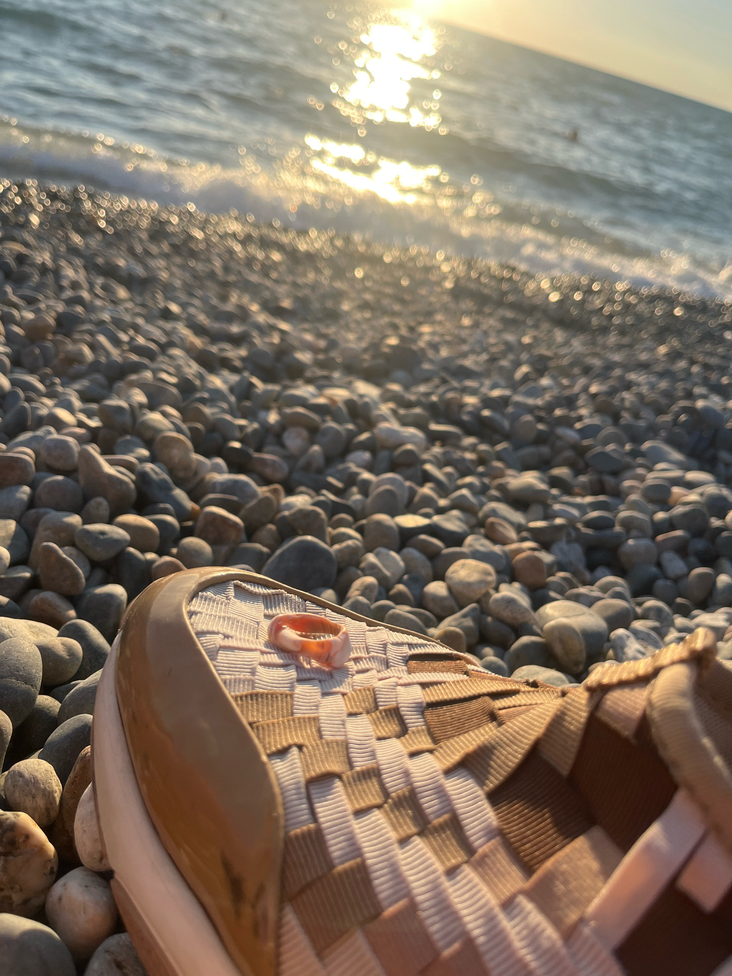 A low-angle, close-up shot captures the toe of a woven sandal resting on a pebble beach. The sandal, in shades of tan and beige, is positioned in the foreground. On top of the sandal, a small, translucent orange ring lies. The background is a blur of the sea and the setting sun, which casts a shimmering reflection on the water's surface. The pebbles are varied in size and color, from light gray to darker hues. The scene is set at the Khostinskiy City beach in Russia.