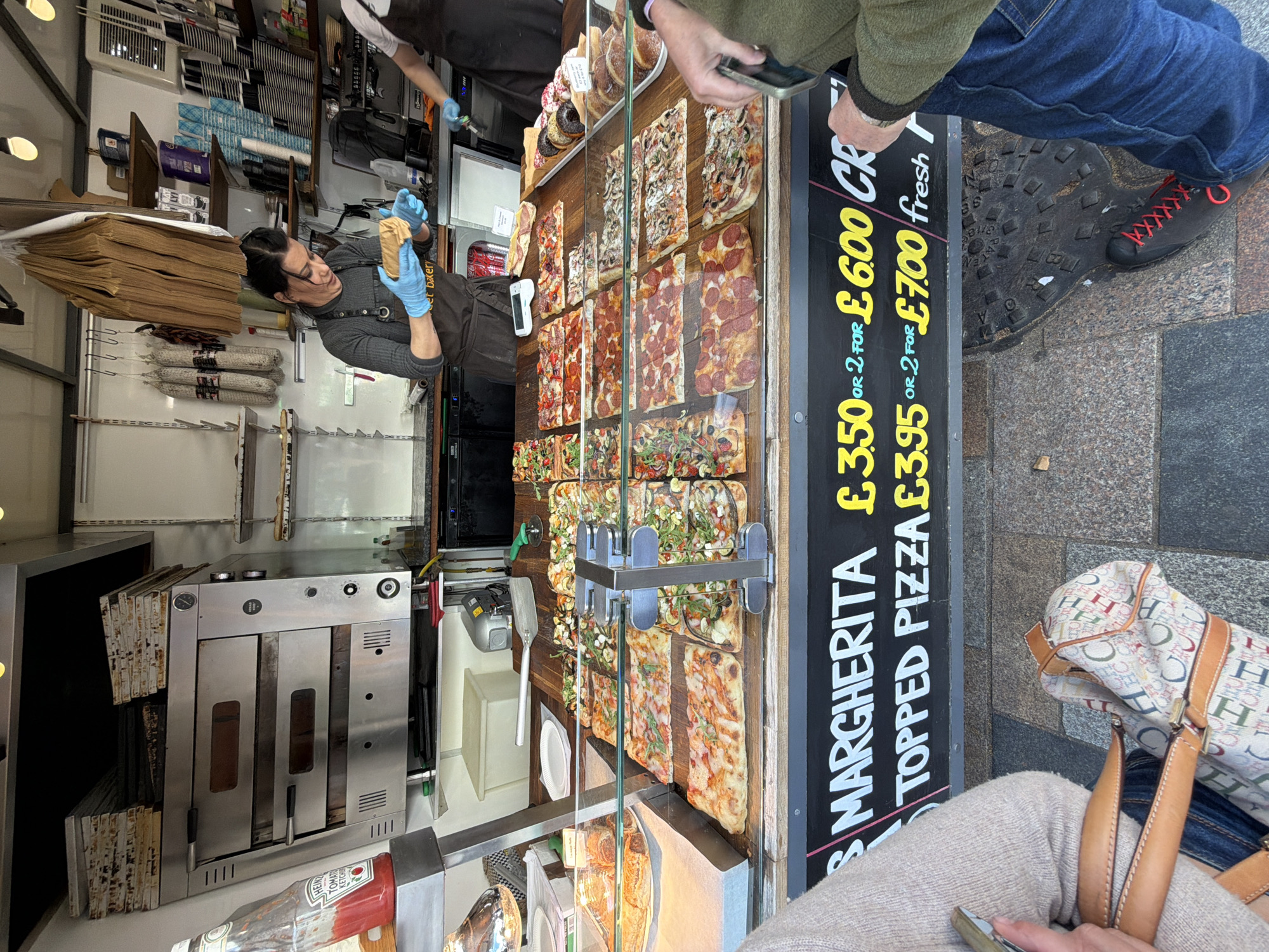 This image captures a lively street food stall or bakery, likely in London, United Kingdom, given the visible currency and general street scene. The perspective is from a customer's viewpoint, looking down and across the counter.
In the center, a food vendor, wearing a dark apron and light blue gloves, is holding a pastry, possibly a croissant or pain au chocolat, appearing to serve a customer. Behind them, stacks of paper bags and some salami hang, while a commercial pizza oven is visible to the left. The stall prominently features a glass display case filled with a variety of rectangular pizzas with different toppings, such as pepperoni, mushrooms, and mixed vegetables. There are also pastries like donuts on display.
A black sign with yellow and white text advertises prices: "MARGHERITA £3.50 OR 2 FOR £6.00" and "TOPPED PIZZA £3.95 OR 2 FOR £7.00 fresh".
Several customers are partially visible. One person's hands are seen holding a smartphone near the counter. Another's legs and a black shoe with red laces are visible on the pavement, standing on a cast-iron drain cover with patterns and the number "9" embossed on it. A third customer's lap and a patterned handbag with "H" monograms are visible in the foreground.
The bright lighting suggests it is daytime. A Heinz Tomato Ketchup bottle is also seen on the counter. The scene conveys a bustling urban environment where food is prepared and sold for takeaway.