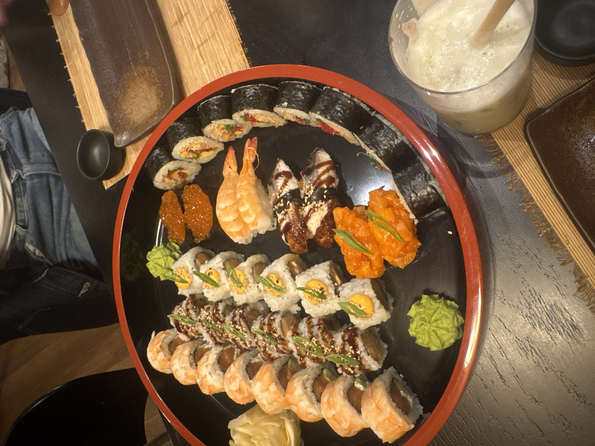 A high-angle, slightly off-center photograph depicts a sushi platter on a dark wooden table, likely in a restaurant in Tbilisi, Georgia. The platter is a round, black tray with a red rim. The sushi arrangement features a variety of types, including maki rolls, nigiri, and potentially temaki or other forms, with fillings such as shrimp, eel, fish roe, and spicy tuna. Condiments such as wasabi and pickled ginger are present. Adjacent to the platter is a tall glass filled with a light-colored beverage and a wooden straw, along with a small black dish and a dark-colored rectangular plate, likely for soy sauce or other dipping sauces. Parts of a bamboo placemat are visible. A portion of a person's lap, clad in denim jeans, is visible in the lower left corner of the frame.