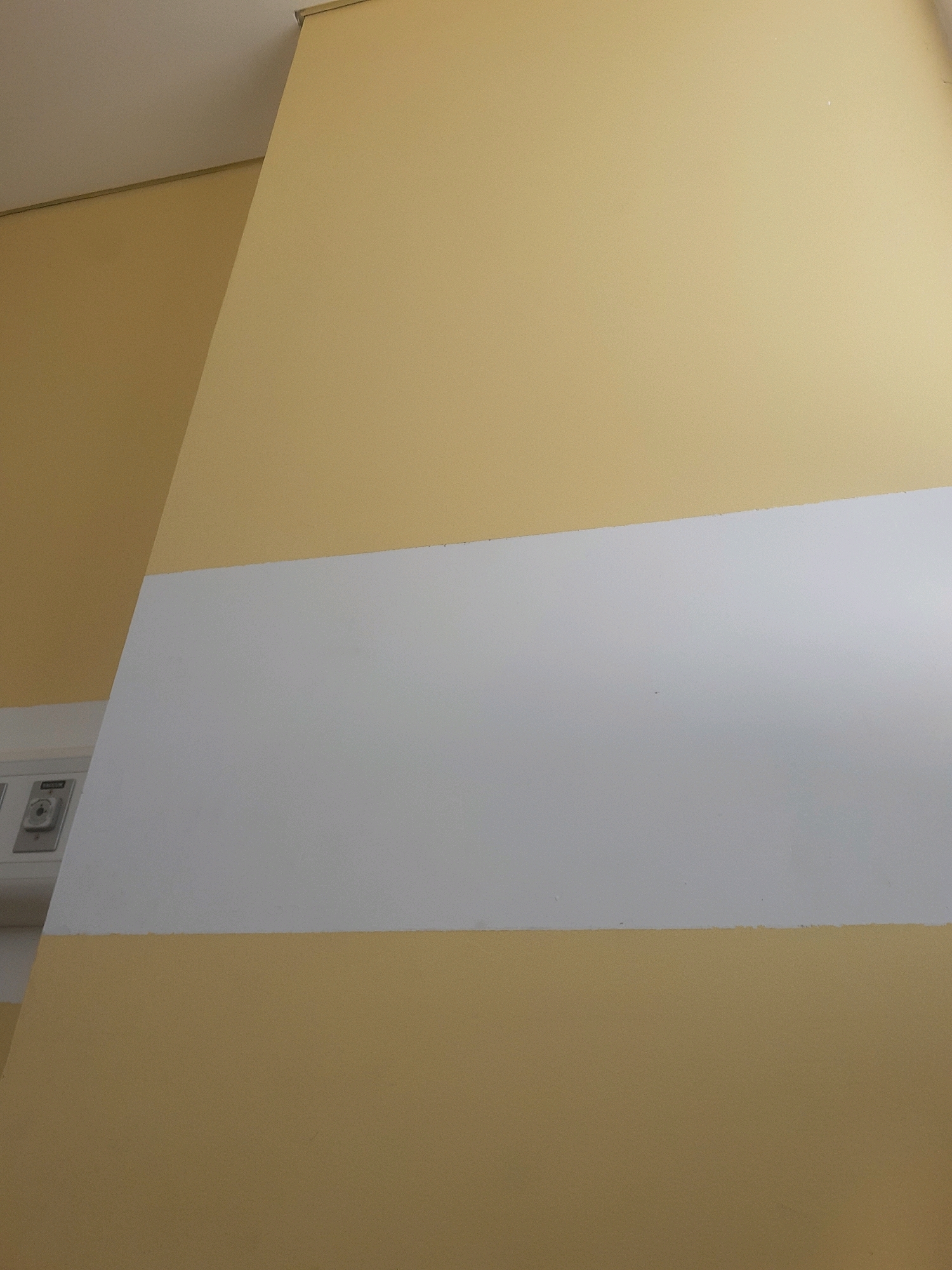 The image shows a close-up of a wall with a horizontal white stripe painted across its surface. The wall is painted in two shades of yellow, with a lighter shade at the top and a slightly darker shade at the bottom. The white stripe is centered on the wall, dividing the two yellow sections. To the left of the image, a portion of a white wall-mounted fixture is visible, featuring a dial and a button, possibly related to medical equipment or utility services. The environment appears to be an interior space, potentially a hospital room or a similar institutional setting. The lighting is soft and diffused. There are no people or animals present in the frame. The image does not provide any specific clues or information that would allow for its location in Sumedang, Indonesia.