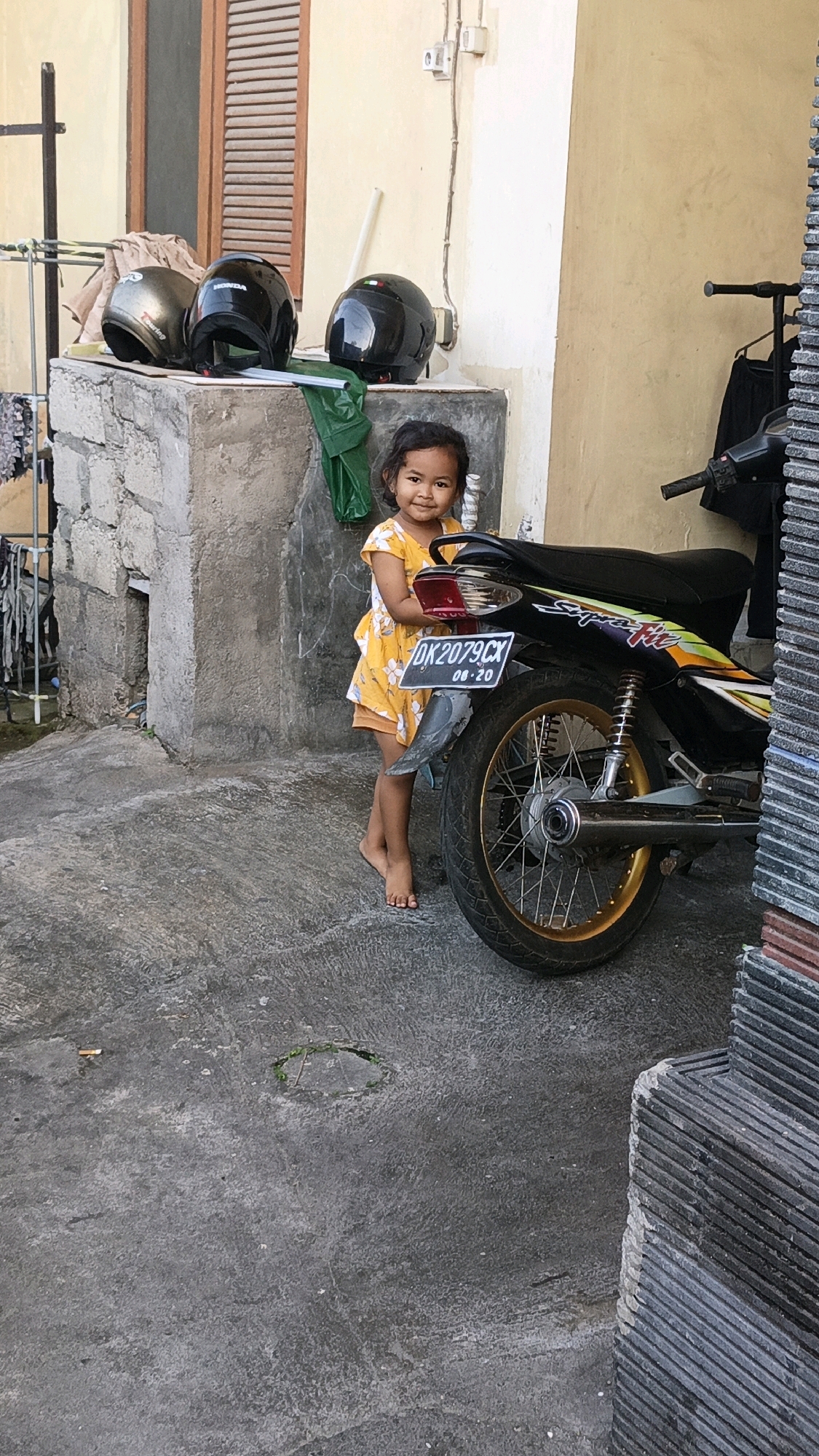 A young girl in a yellow dress stands next to a motorcycle in a concrete courtyard. The girl is looking at the camera and smiling. The motorcycle is black with gold accents and has a license plate that reads "DK 2079 CX 08-20". Behind the girl, there are three helmets on a concrete ledge. The environment appears to be an outdoor residential area in North Kuta, Indonesia, with a wall and a window visible in the background.