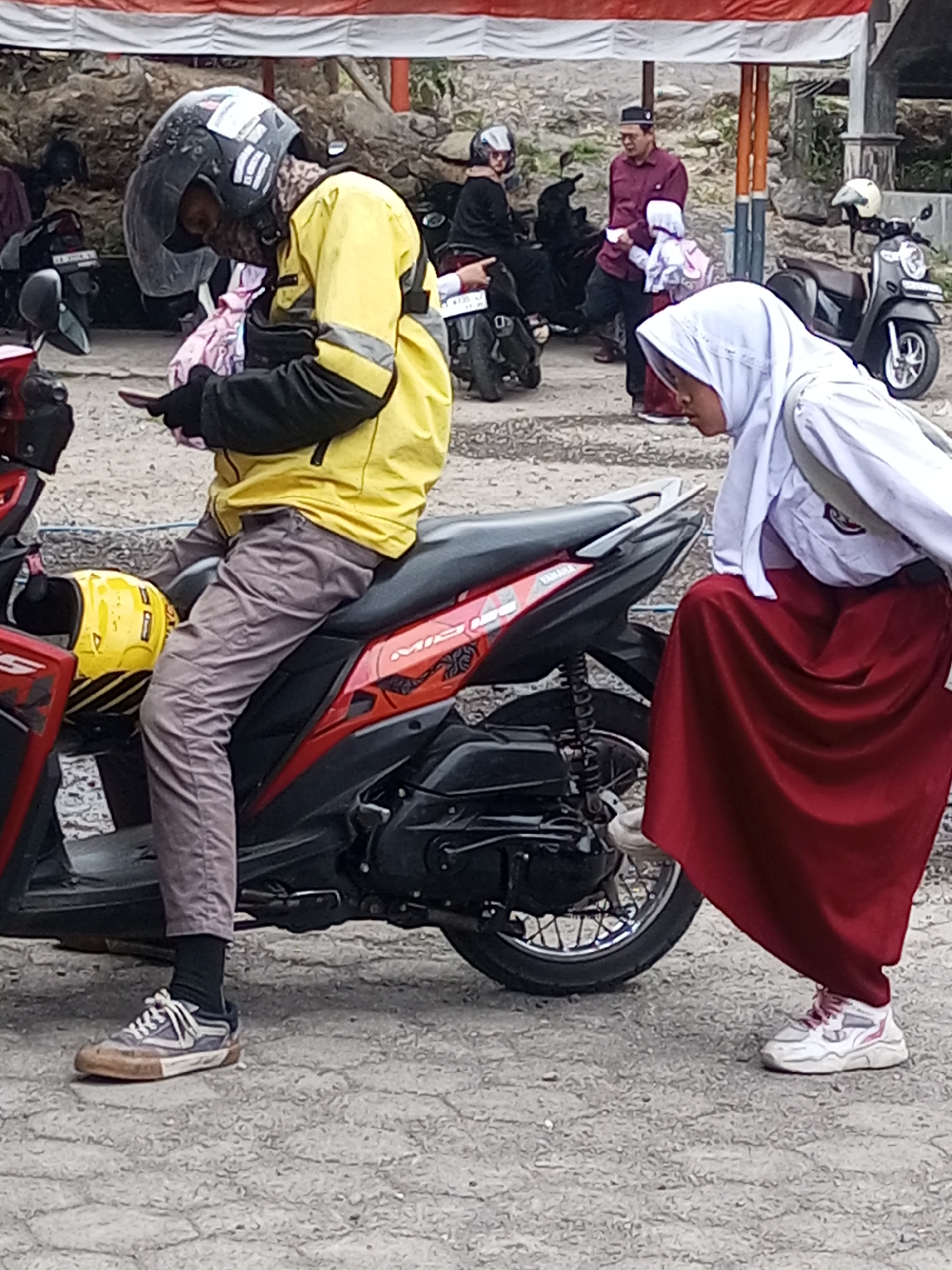 In Krasak, Indonesia, a person wearing a yellow jacket and a helmet sits on a red and black motorcycle. They are looking down at their phone. To the right, a person wearing a white hijab and a red skirt stands next to the motorcycle, with their foot resting on the bike's rear wheel. In the background, several other motorcycles are parked, and some people are visible, including a man in a maroon shirt and a child in a pink backpack. The scene appears to be outdoors, possibly in a parking area or on a street, with a textured gray ground surface.