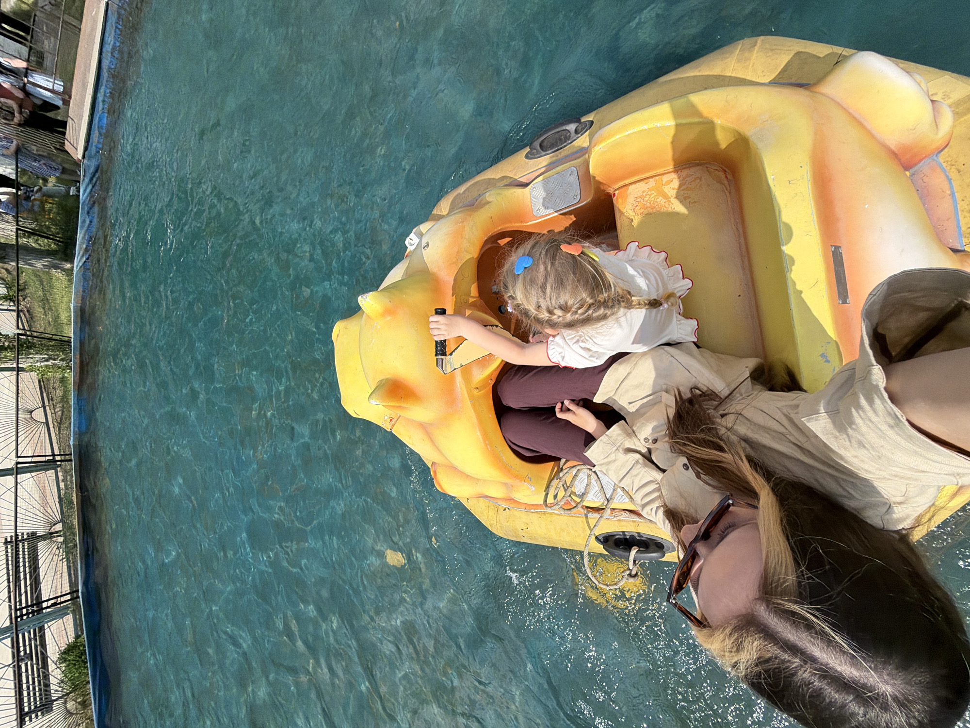 A slightly rotated, high-angle, outdoor shot depicts a young girl and an adult in a yellow, animal-shaped paddle boat on a body of water.
The girl is positioned in the front seat, holding a black steering handle. She has blonde, braided hair adorned with blue and orange hair clips. She wears a white, ruffled-sleeve shirt and dark pants.
The adult is seated beside and behind the girl. The adult has long, brown hair with blonde streaks and wears sunglasses and a tan jacket.
The boat is a bright yellow color and appears to be shaped like a stylized animal. It is floating on a body of water that appears blue-green in color.
Along the left edge of the frame, there is a metal fence and a walkway.
The location is identified as Kamyshin, Russia.