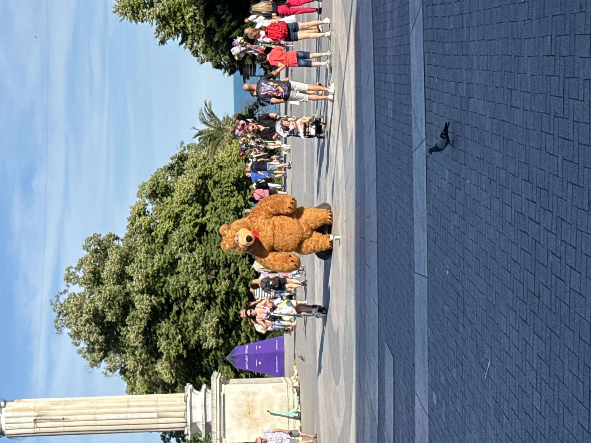 An adult person in a brown bear mascot costume stands on a paved plaza in front of a large green tree. The bear is facing left, and its paws are slightly apart. To the left of the bear, a person rides a scooter. In the background, other people are walking, and there are balloons and a purple pyramid-shaped structure. The sky is blue with some wispy clouds. In the foreground, a pigeon walks on the paved ground.
