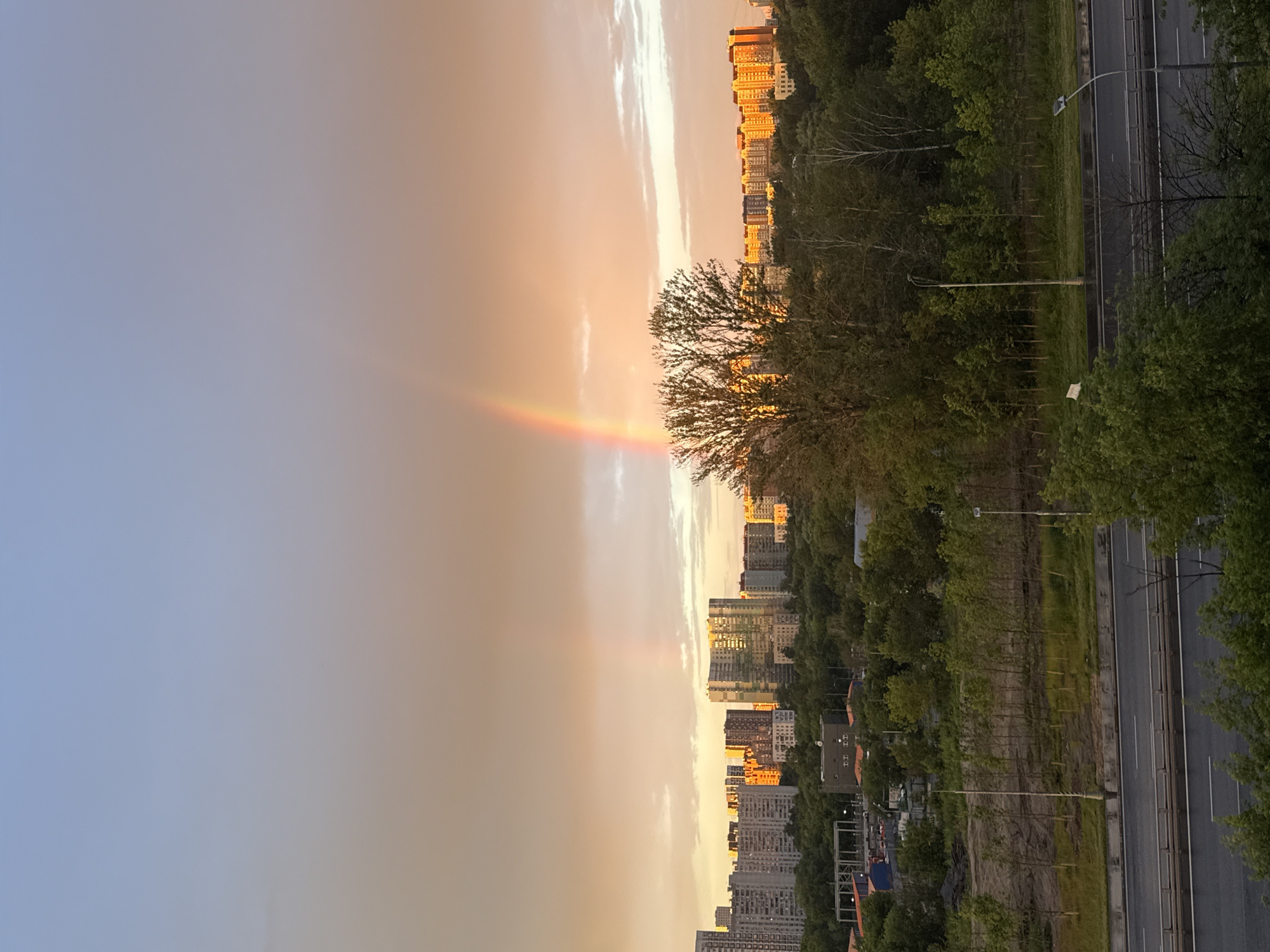 The image depicts a view of Moscow, Russia, during a sunset with a rainbow. In the background, a cityscape is visible with numerous apartment buildings. The sky is illuminated by warm sunset hues, ranging from light blue at the top to orange and yellow near the horizon. A prominent rainbow arches across the sky, with its vibrant colors clearly visible against the cloudy backdrop. In the mid-ground, a dense green forest separates the cityscape from a highway in the foreground. The highway appears to be wet, suggesting recent rain. Trees and streetlights are also present along the highway. The overall scene conveys a peaceful and picturesque moment in an urban environment.