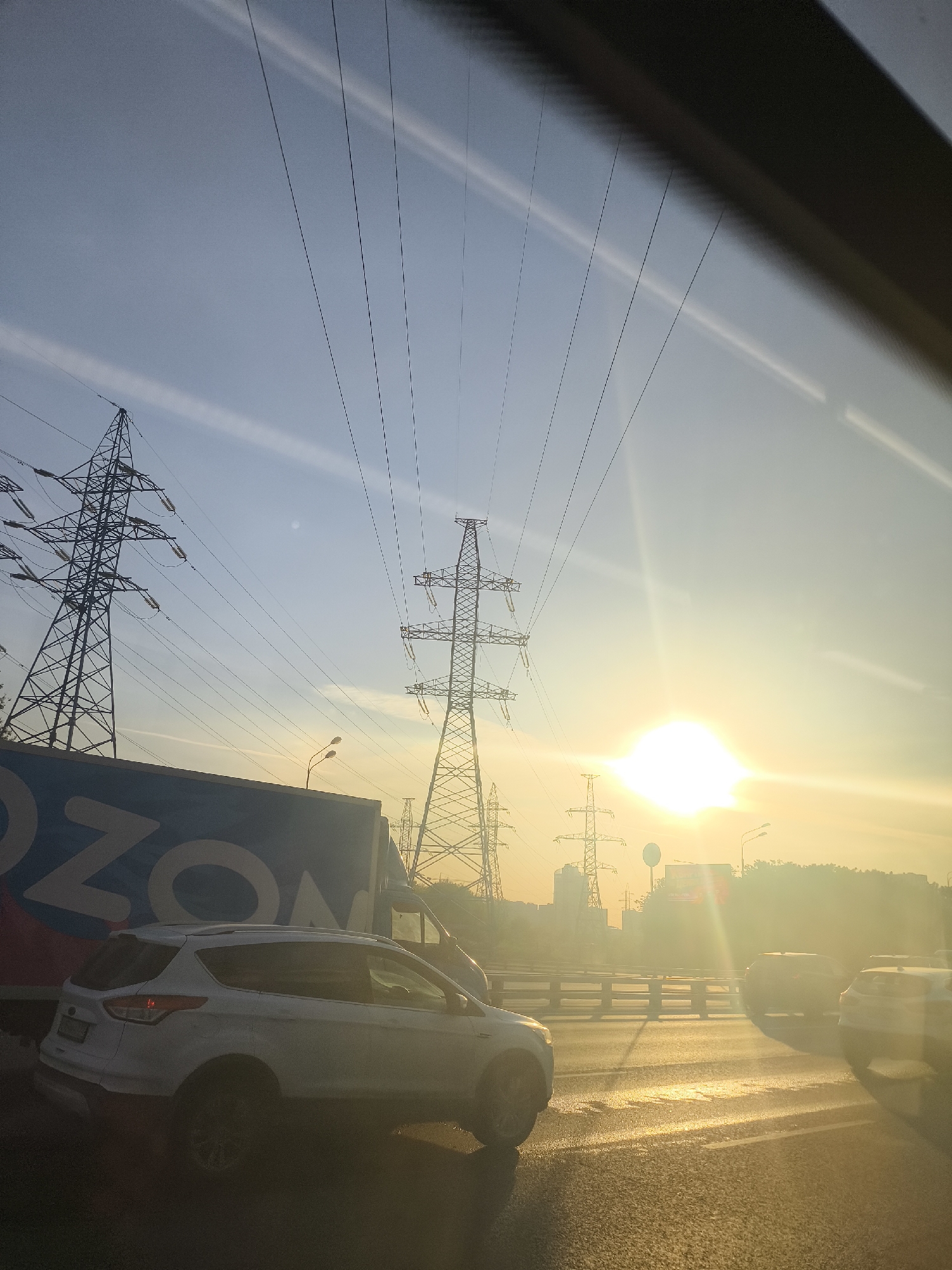 The image depicts a roadside scene, likely taken from within a moving vehicle, as evidenced by the blurred foreground suggesting a window or windshield frame. The prominent feature is a series of electrical power transmission towers against a sky illuminated by the sun, which casts a warm, golden glow across the horizon. A large truck, partially visible on the left, bears the "OZON" logo, indicating a logistics or delivery service. A white SUV is parked in front of the truck. Other vehicles can be seen on the road ahead. The setting appears to be an urban or suburban environment, but specific details about the exact location are not clear. The time of day appears to be morning or late afternoon based on the lighting.