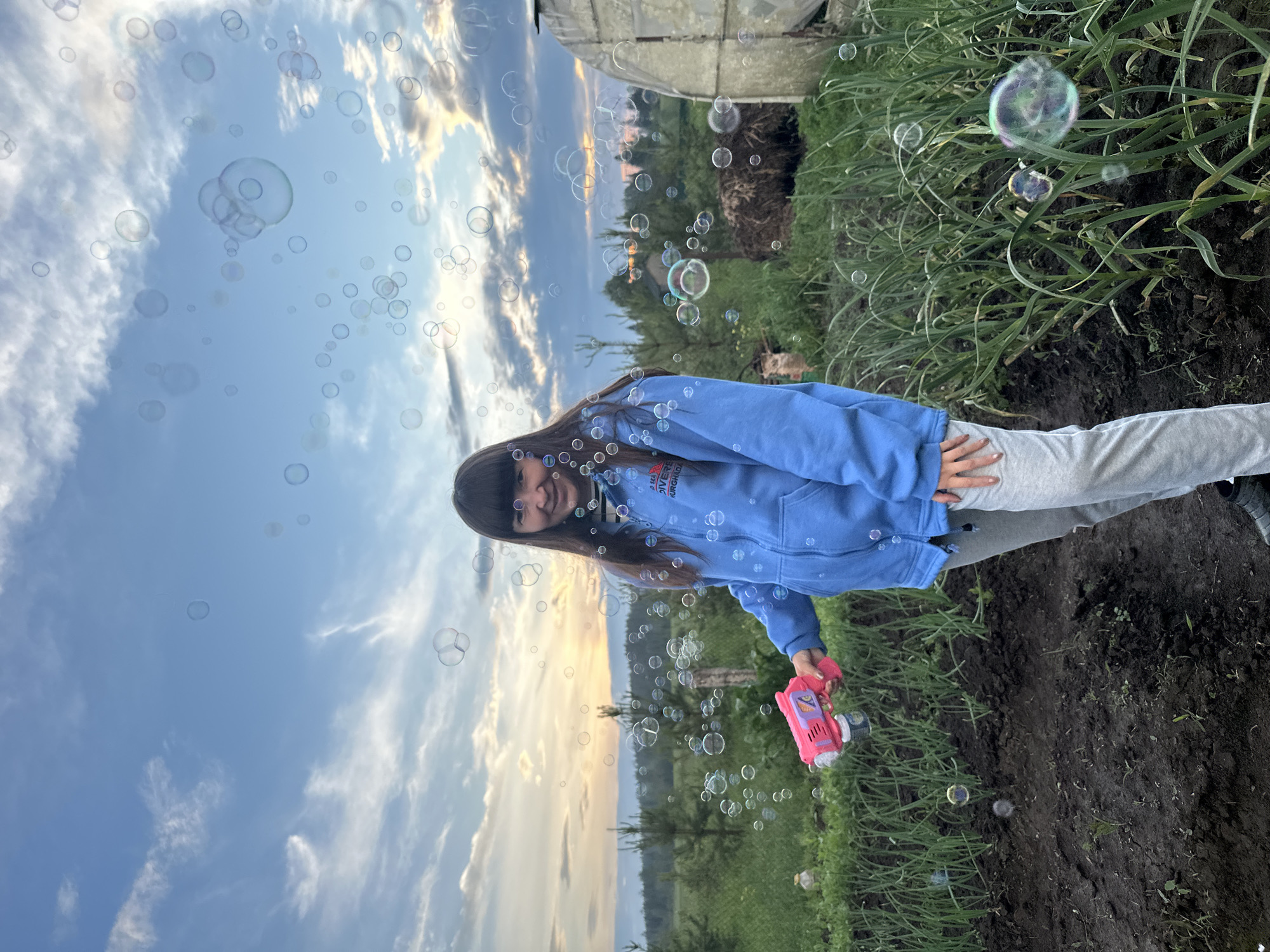 An outdoor, medium shot captures a young female with dark hair and bangs, wearing a blue hooded jacket and gray sweatpants, standing in a garden. She is holding a pink bubble gun in her right hand, pointed towards the left, and appears to be blowing bubbles. The background features a greenhouse on the right, rows of green plants, and a forest in the distance under a cloudy sky with sunset hues. The location is identified as Buranovo, Russia.