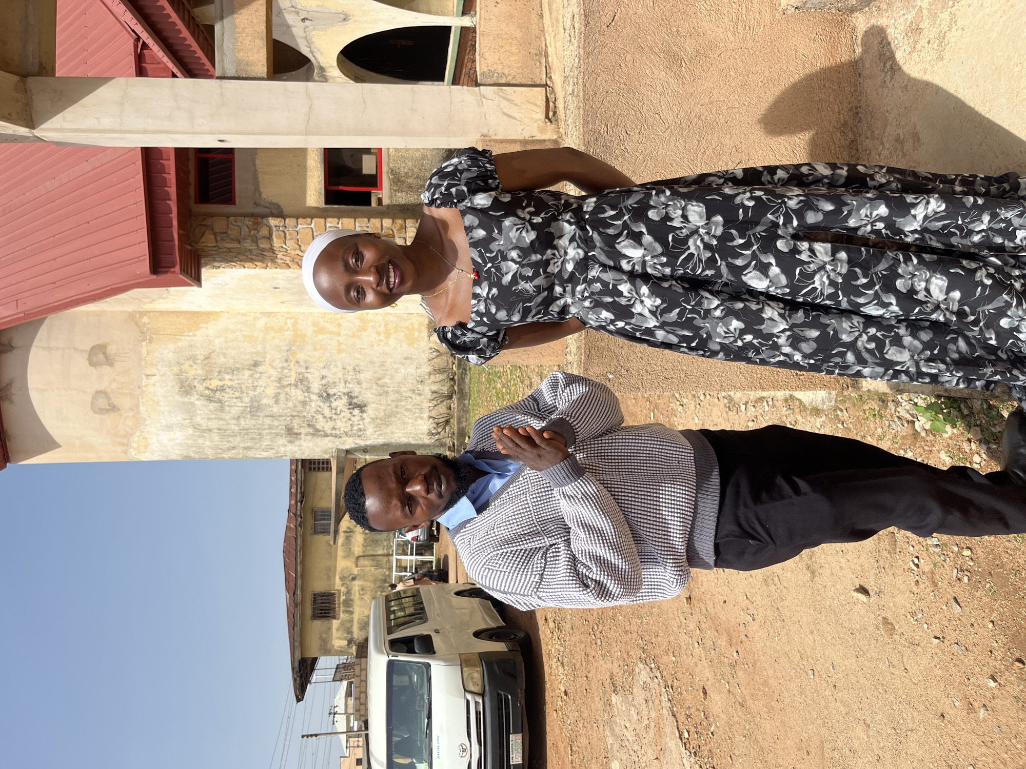 In the city of Bukuru, Nigeria, a man and a woman stand outside a building with a red-tiled roof. The man, on the left, is wearing a light blue collared shirt and a patterned sweater over black pants. He has his hands clasped in front of him. The woman, on the right, is wearing a black and white floral print dress and a white headscarf. She is smiling at the camera. Behind them, a white van is parked on a dirt road. The building appears to be a place of worship, possibly a church, with arched doorways and rough-textured walls. The sky is clear and blue.