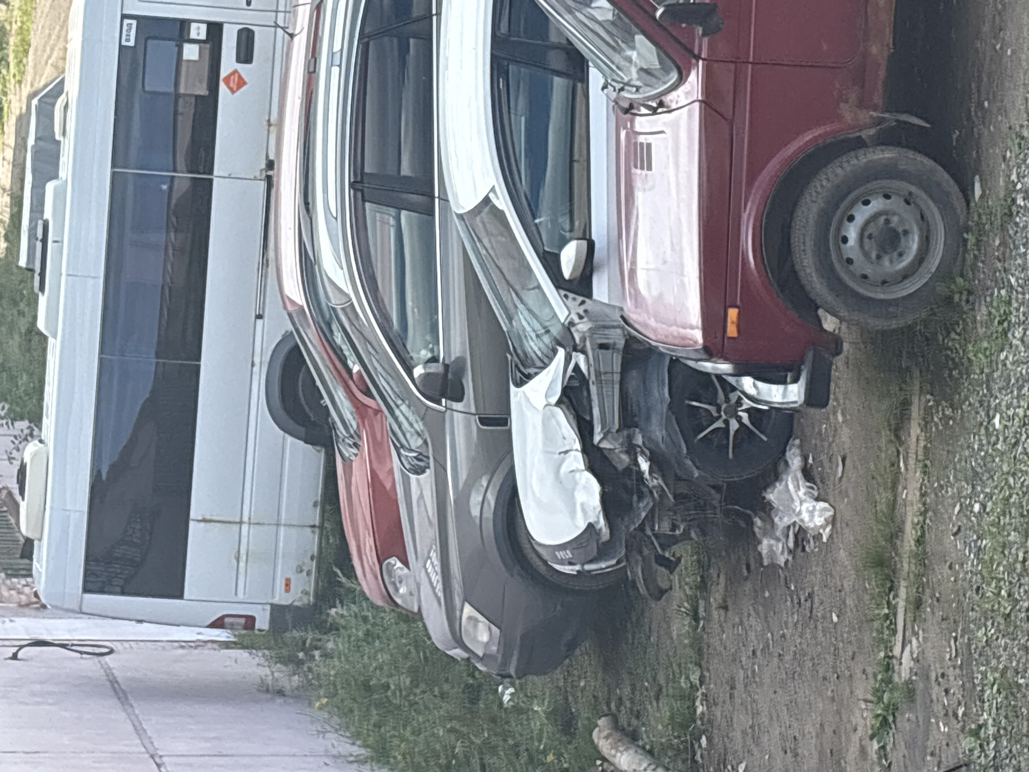 The image depicts a lineup of several cars parked on a gravelly surface next to a concrete wall. In the background, a white minivan is visible. In the foreground, a row of cars includes a dark gray sedan, a red sedan, and a damaged white vehicle with its front end crushed. A red SUV or hatchback is parked in the front right. The overall scene suggests an informal parking area or a junkyard, possibly in an urban or suburban setting, potentially Noviy, Russia, given the context. The environment appears somewhat neglected, with overgrown grass and debris on the ground.