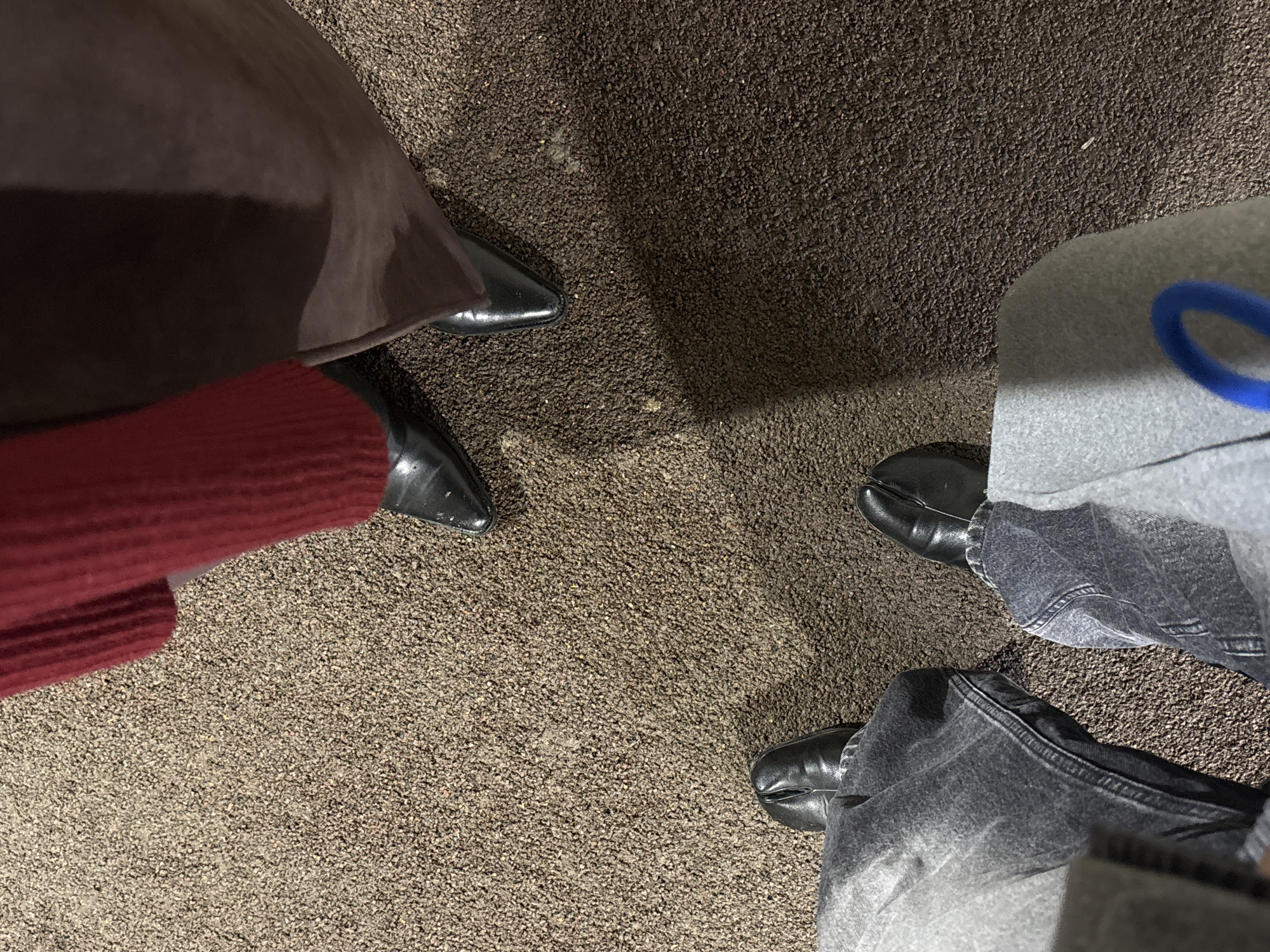 The image shows a close-up of three pairs of feet on a brown carpet. The first pair is wearing black pointed-toe shoes, partly covered by a brown garment (possibly a skirt or coat) and a red garment (possibly a sweater). The second pair is wearing black shoes and faded black jeans. The third pair is wearing black shoes and gray jeans, and has a blue object (possibly a bag handle) and a gray object (possibly a container) nearby. The environment is a brown, textured carpet, typical for indoor flooring. The location is unknown but the prompt suggests Perm, Russia. There are no clear indications in the image that would confirm this.