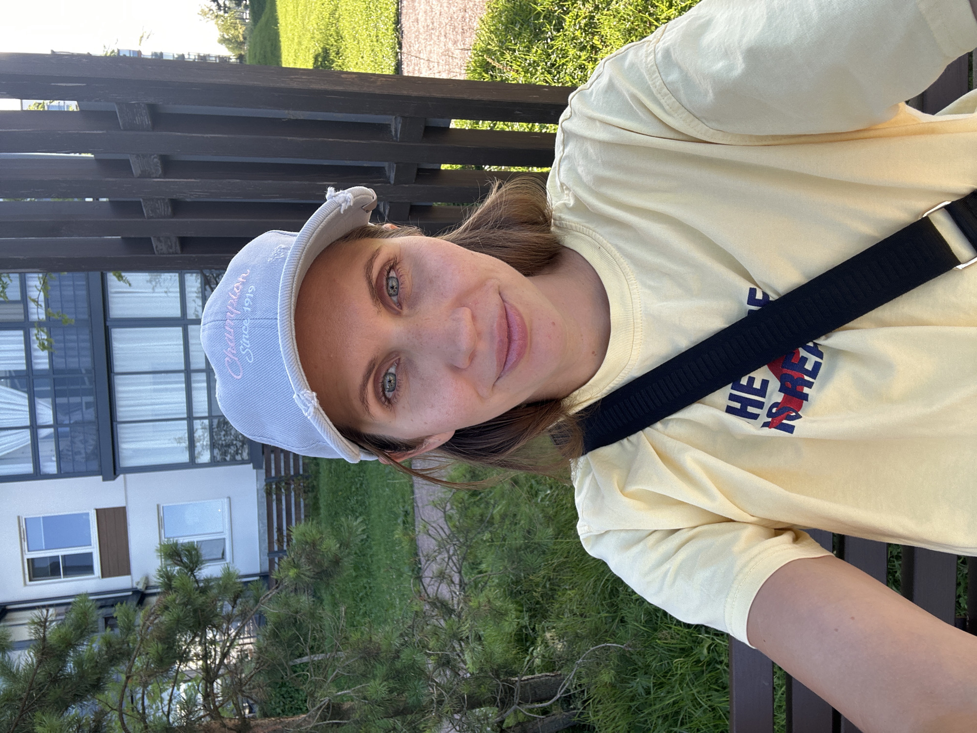 A woman with blonde hair, wearing a light blue cap and a yellow t-shirt, takes a selfie outdoors. The cap has "Champion Since 1919" embroidered on it. She has a black strap across her chest. In the background, there is a modern building with windows and balconies, green grass, a pathway, a pine tree, and a dark wooden fence. The setting appears to be a residential area in Vsevolozhsk, Russia.
