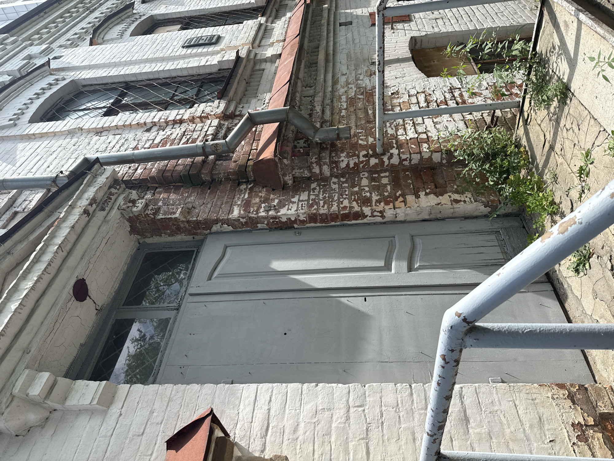 This is a photograph of the exterior of an old, two-story brick building with white painted facade, located in the city of Khabarovsk, Russia. The building appears to be in a state of disrepair, with peeling paint and weathered brickwork. A large, grey wooden door with a single pane window at the top is prominently featured on the left side of the frame. The door is weathered and shows signs of age. To the right of the door, the brick wall is exposed, revealing red brick and mortar, with sections of white paint flaking off. A metal downspout runs vertically down the side of the building. Above the door, a decorative white molding with cracks is visible. The building has arched windows with diamond-shaped metal grates. A rusty metal railing runs along the front of the building. Small green plants are growing through cracks in the pavement and around the base of the building, indicating neglect. The lighting suggests it is a sunny day, casting shadows on the door and pavement.