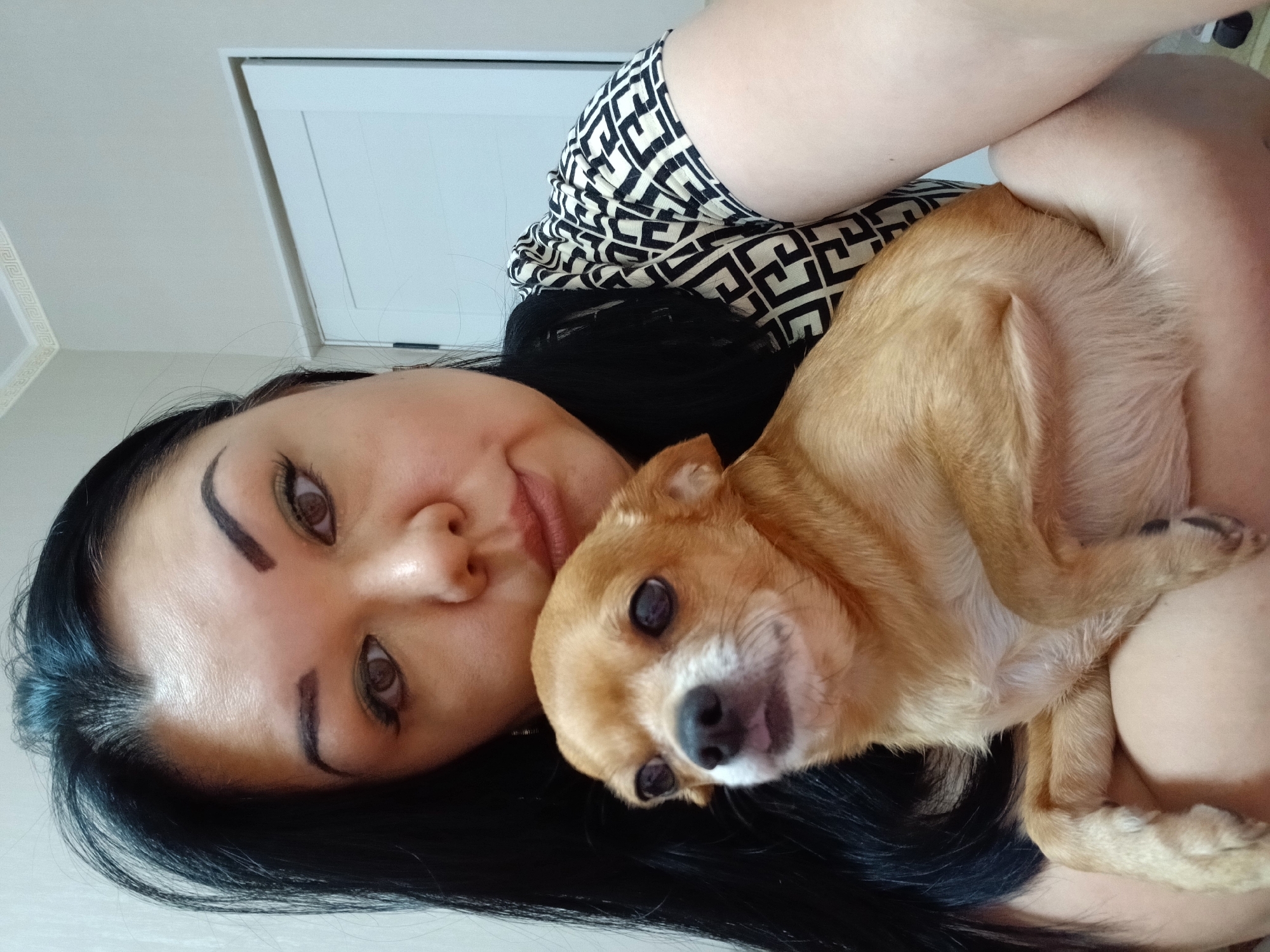 A close-up, waist-up selfie shows a woman with long, dark hair and defined eyebrows holding a small, light brown chihuahua. The woman is wearing a black and white patterned top. The chihuahua is looking towards the left of the frame with its tongue slightly out. The background appears to be an indoor setting with a white door and a patterned border along the top of the wall.