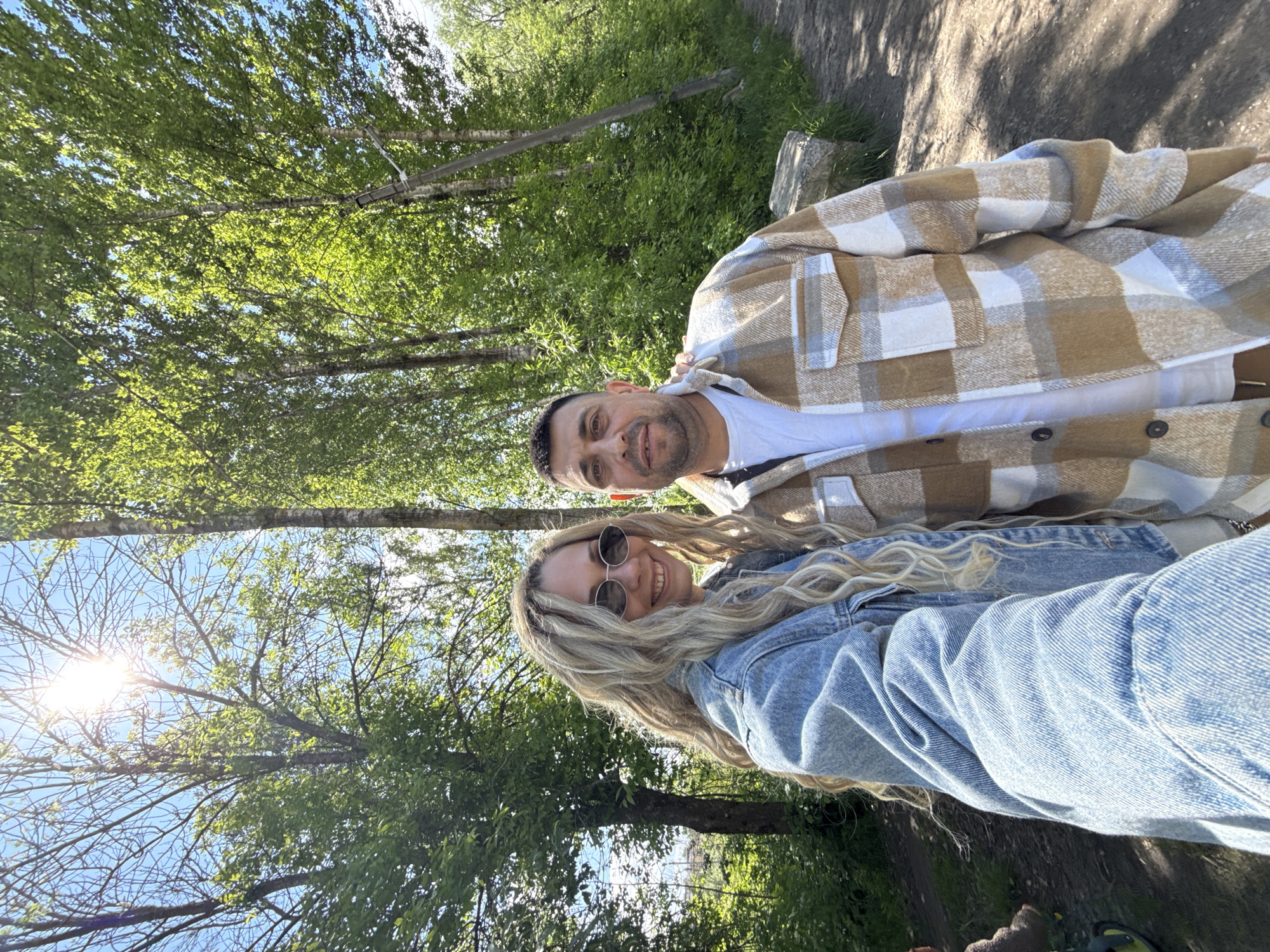The image shows a man and a woman posing for a selfie in an outdoor, natural setting. The woman is wearing a light blue denim jacket and sunglasses, with long blonde hair. The man is wearing a white t-shirt and a brown and white checkered jacket. They appear to be smiling. Behind them, there is a background of trees with green leaves, suggesting a park or wooded area. A bright sun is visible through the trees. The ground appears to be a path or trail with some grass and foliage. It is not possible to confirm whether the location is Rusanovo, Russia based on the provided information.