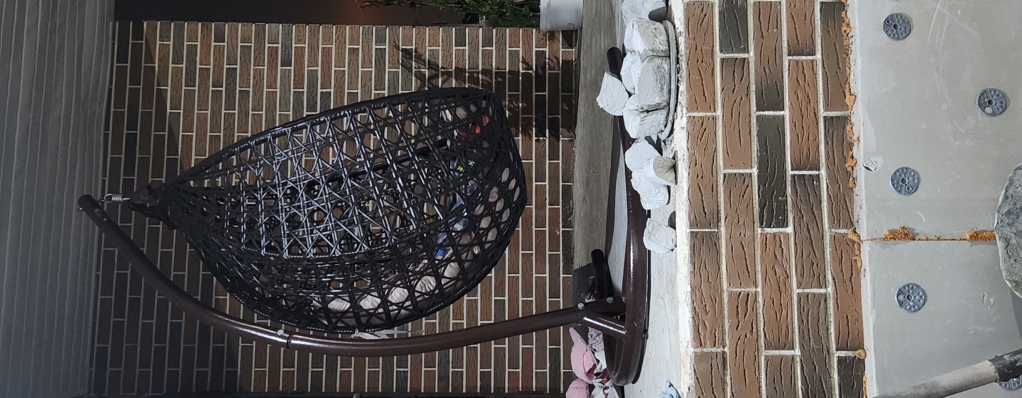 A hanging basket chair made of dark woven material is positioned on a concrete porch in front of a brick wall. The chair is empty, but appears to have some items inside, possibly cushions or blankets, and a few plastic bottles. A brown metal stand supports the chair. Scattered on the porch in front of the chair are several white, irregularly shaped objects, which appear to be broken pieces of plaster or concrete. To the right of the chair, a small potted plant is visible. The bottom of the image shows a section of a building under construction with exposed insulation and what appears to be expanding foam. The scene is dimly lit, suggesting it is either dusk or nighttime.