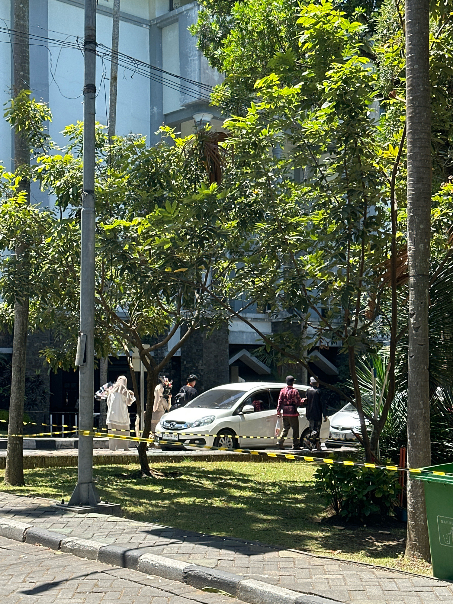 In Malang, Indonesia, a street scene unfolds under bright sunlight. A white car, identified by license plate S 1524 DE, is parked on the side of the road. Yellow caution tape is visible, creating a boundary around the immediate area. Several individuals are present. One person, wearing a white hijab and a long white garment, stands near the car. Another person, also in a light-colored hijab and garment, holds a bouquet of flowers. Two other individuals, both male, are also near the car. One is dressed in a red and black patterned shirt and dark pants, while the other wears a dark shirt and pants. The background features a building with a light-colored facade and dark windows, surrounded by lush green trees and vegetation. A gray lamppost stands prominently in the foreground, with electrical wires running across it. A green trash bin is visible on the right side of the image. The ground is paved with interlocking bricks and concrete curbs.