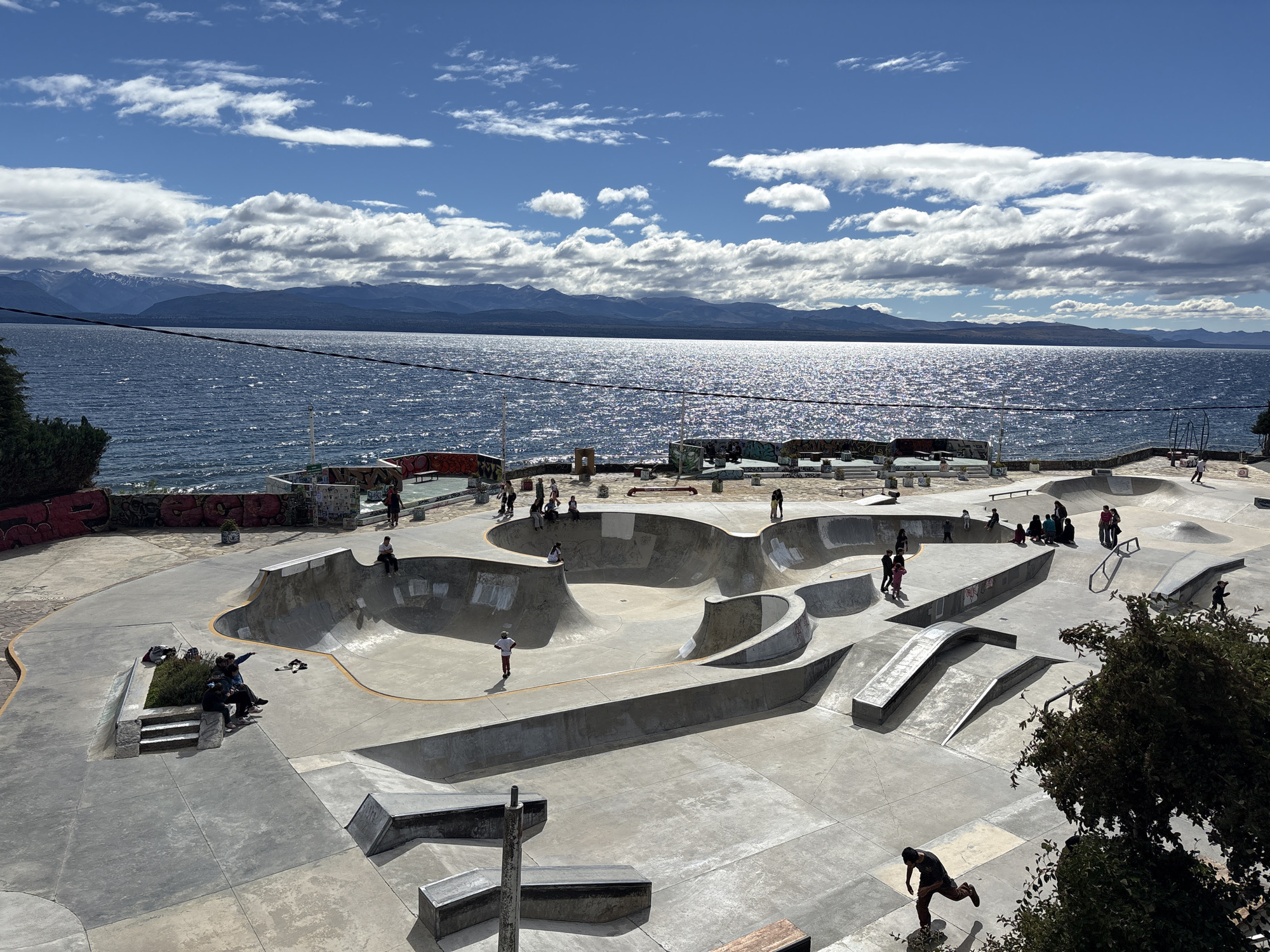 The image depicts a skatepark located in San Carlos de Bariloche, Argentina, overlooking a large body of water (likely a lake) with mountains visible in the background. The skatepark is made of concrete and features a variety of ramps, bowls, rails, and other obstacles. Several people are present, some actively skateboarding and others observing or socializing. The sky is partly cloudy, with the sun reflecting off the water's surface. The area surrounding the skatepark includes trees, decorative blocks, and some painted structures.