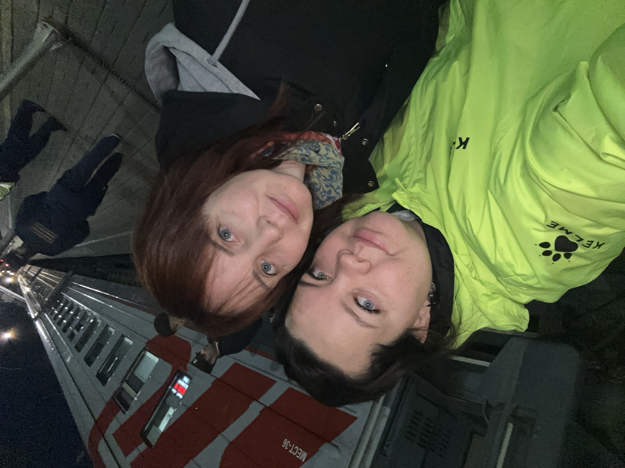 Two women pose for a selfie. The woman on the left has red hair, a black jacket, and a blue and white patterned scarf. The woman on the right has dark hair and is wearing a neon green jacket with "Kelme" and a paw print logo. In the background is a passenger train car with red stripes. The train station appears to be in Izhevsk, Russia, based on the Russian lettering visible on the train car: "МЕСТ-36". Other people are visible in the background, including a person walking on what appears to be a covered walkway or bridge. The picture appears to be taken at night.
