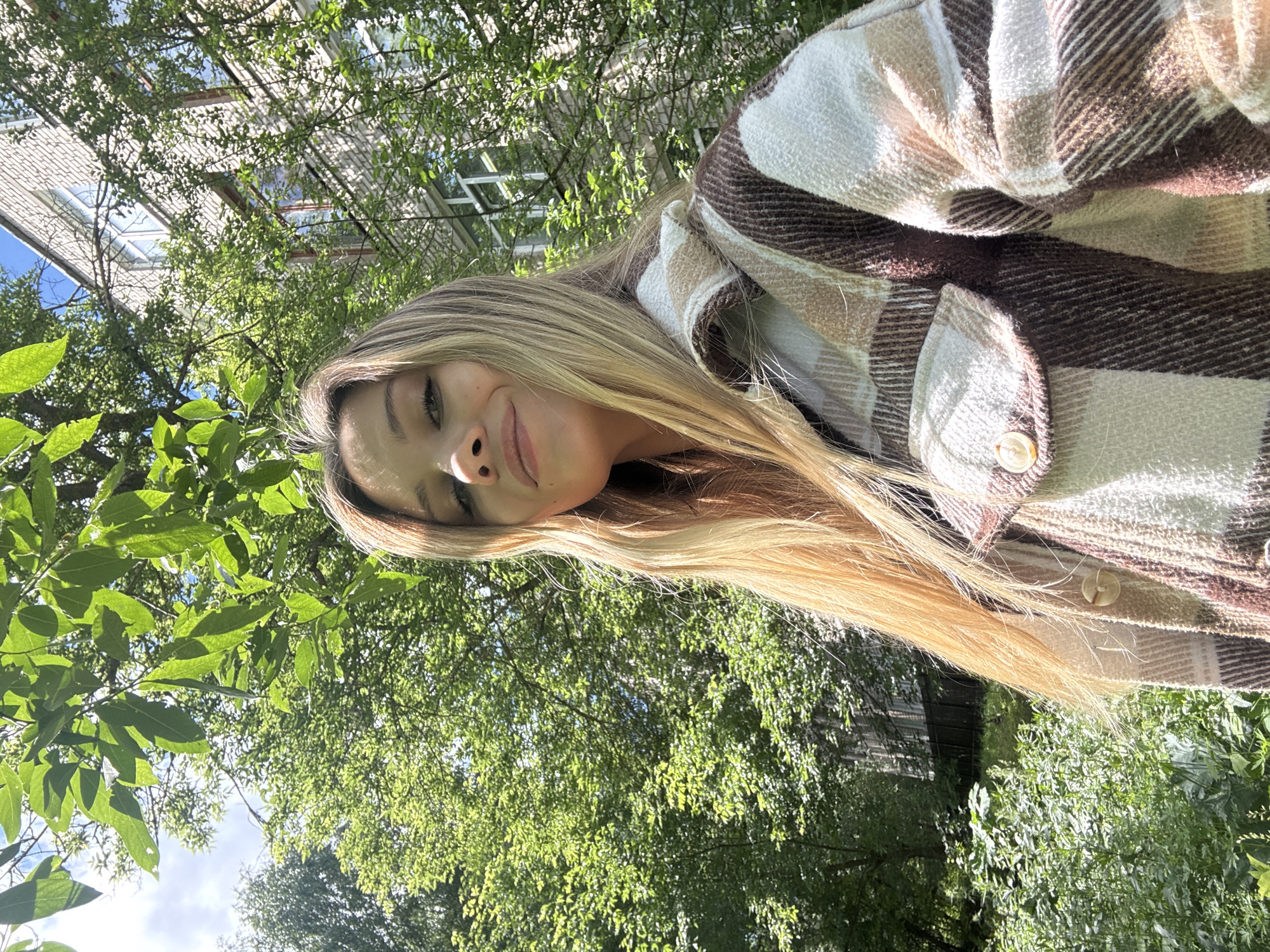 An outdoor, waist-up selfie shows a young woman with long, blonde hair and a plaid jacket. She is smiling and looking at the camera. Behind her, there are trees and a building. The building appears to be a multi-story apartment building made of light-colored brick. The trees are full of green leaves, suggesting it is summer or late spring. The sky is blue with some white clouds. The scene is set in a residential area, likely in the city of Chaikovsky, Russia, given the architectural style of the building and the surrounding vegetation.