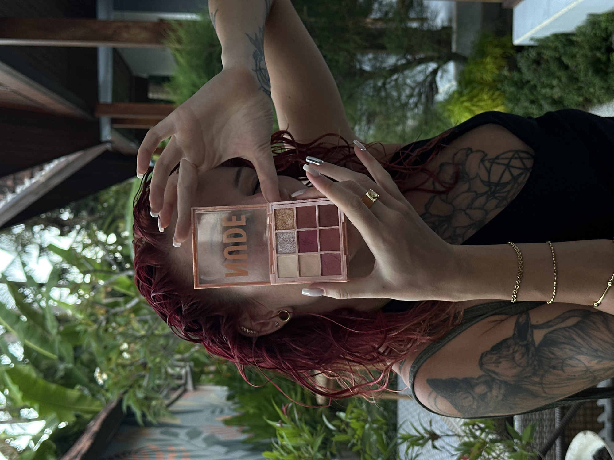 A person with red hair and tattoos is holding a "MODE" eyeshadow palette in their hands. The person's right hand has a gold ring on one of the fingers. The background features green plants and a building structure. The location is Nusa Penida, Indonesia.