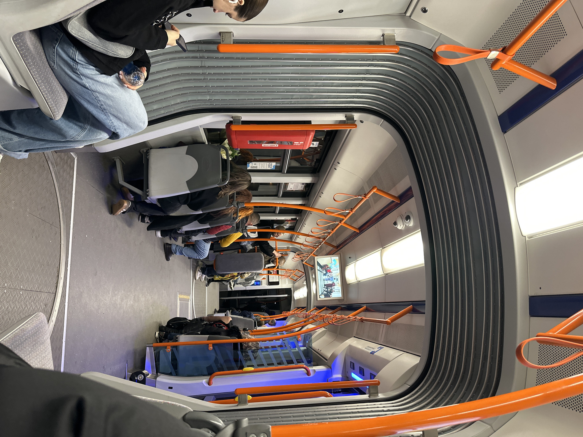 The image captures the interior of a train car, seemingly in motion, with passengers seated along both sides. The train's interior is dominated by rows of seats and a curved wall with a distinctive gray ribbed design. Orange handrails and loops are visible along the ceiling and sides, aiding standing passengers. Seated passengers are visible, with varying demographics. The floor appears to be a textured metal, and overhead lighting is present. A screen mounted on the wall displays content, potentially advertisements or informational displays. Based on the distinct aesthetic of the train's interior, the location is likely within the city of Sofia, Bulgaria.