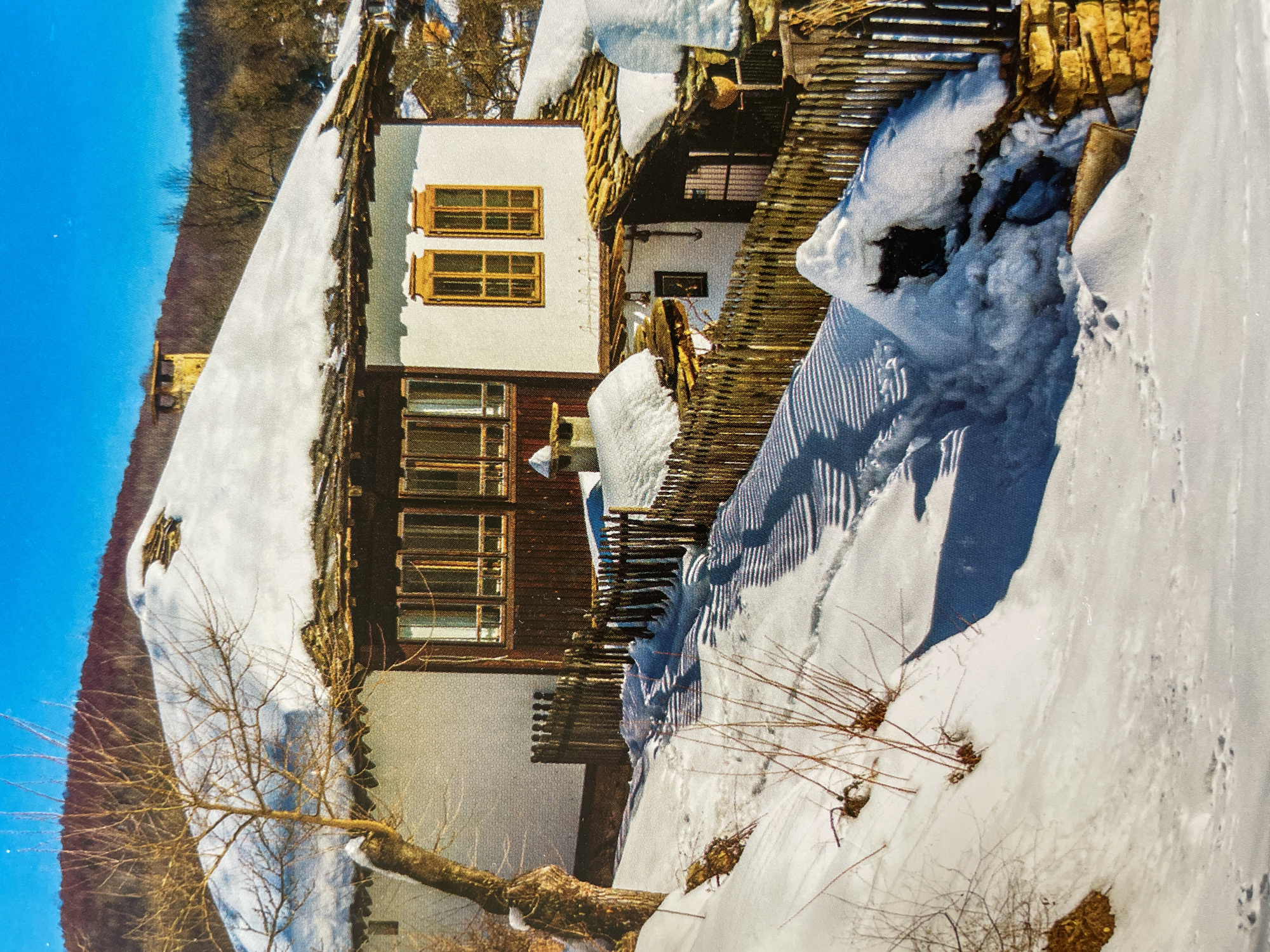 The media file captures a serene winter landscape, prominently featuring a traditional house heavily blanketed in snow under a clear blue sky. The image is oriented with a significant tilt.
The main subject is a two-story house with a distinct architectural style. Its upper section is painted white, while the lower part features dark wooden siding. Multiple windows are visible, characterized by bright, possibly yellow or light brown, wooden frames and shutters. The roof of the house is entirely covered in a thick layer of white snow. A rustic wooden fence, also partially submerged and covered in snow, runs along the side of the house, indicating a property boundary.
The ground in the foreground and around the house is covered in deep, undisturbed snow, casting long shadows that suggest bright sunlight. Bare, deciduous tree branches and sparse, dry bushes emerge from the snow, adding texture to the wintry scene. In the background, rolling hills or mountains, appearing dormant and possibly covered with thin trees, rise against a vibrant, cloudless blue sky.
There are no people or explicit activities depicted. The scene conveys a quiet, cold winter day. No visible text is present in the image. The general setting suggests a rural or mountainous environment, consistent with winter scenes found in regions like Sofia, Bulgaria, as per the provided context.