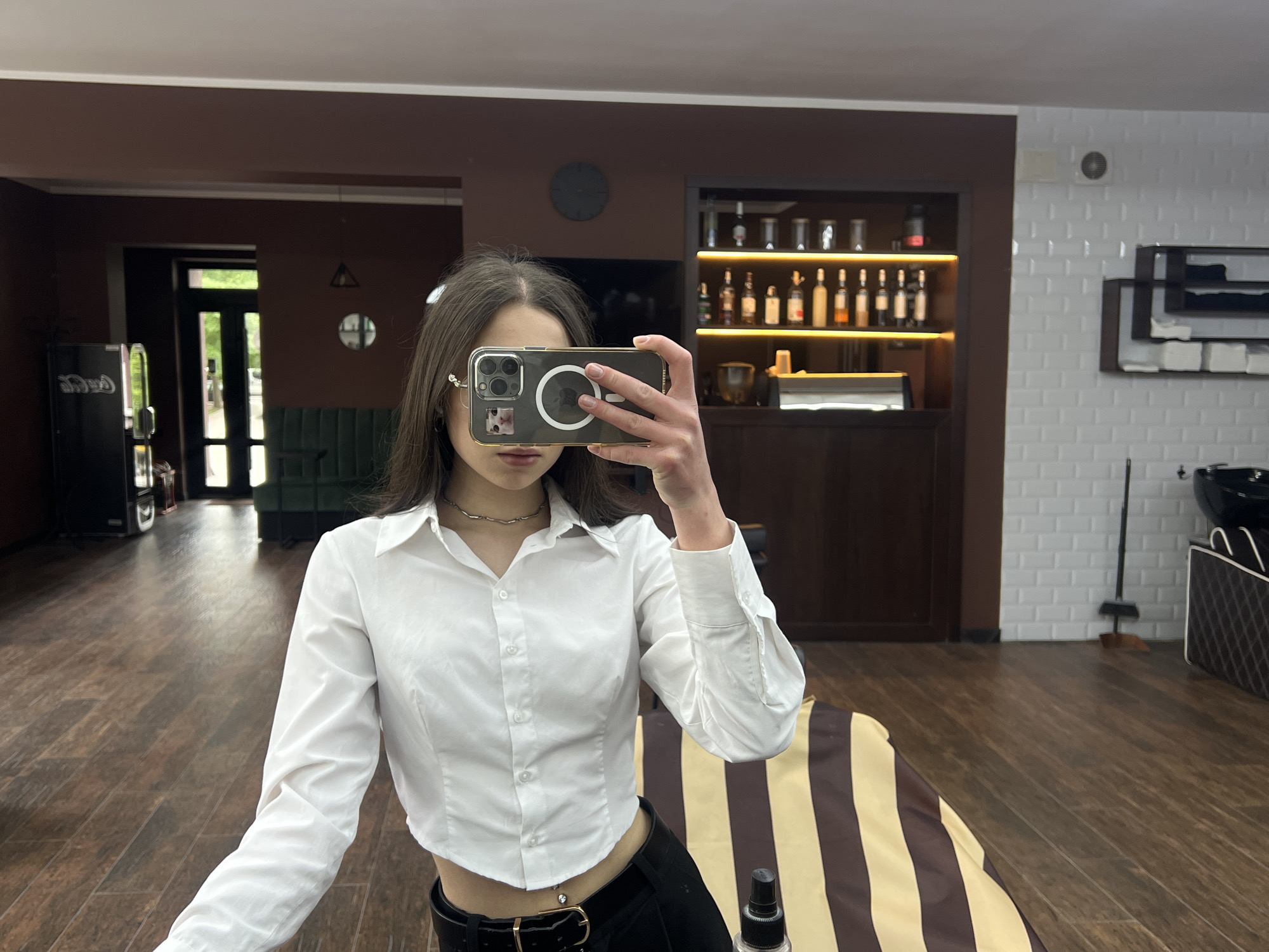 A young woman takes a selfie in what appears to be a hair salon, likely in Ternopil, Ukraine. She is wearing a white cropped button-up shirt, black pants with a belt, and a silver chain necklace. She holds up an iPhone, obscuring her face. The salon features brown and white tiled walls, wooden floors, a bar with illuminated shelves holding bottles, a brown bench, a sink, and a barber's chair covered in a brown and yellow striped cape. A refrigerator is visible through the open doorway in the background.