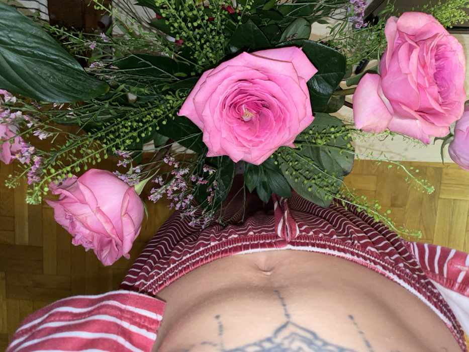 The scene is set indoors in Boreti, Montenegro. The left side of the frame features a partial human torso, displaying a bare abdomen with a central navel. Dark, patterned tattoos are visible on the upper left hip area. The individual is wearing red and white striped loungewear, characterized by a waistband with a wavy red and white pattern. The right side of the image is dominated by a floral arrangement. This arrangement consists of multiple vibrant pink roses, presented in various stages of bloom, interspersed with diverse green foliage. The greenery includes broad dark leaves, delicate sprays of small green berries or buds, and sparse clusters of tiny light purple floral accents. The background reveals a light brown parquet wood floor.