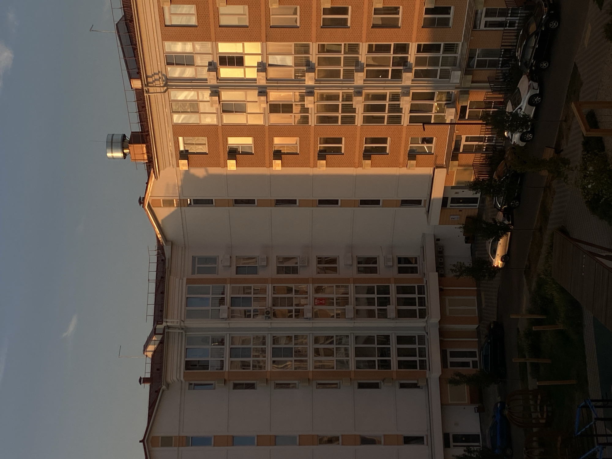 The image captures a view of apartment buildings in Vishnyaki, Russia, during what appears to be sunset or sunrise, given the warm, golden light illuminating the brick facade of one building. The sky is a gradient of soft blue and grey, with a faint, wispy cloud formation.
Two multi-story apartment buildings dominate the frame. The building on the left has a white facade with orange trim around the windows and balconies. The building on the right features a prominent brick exterior, reflecting the warm light. Both buildings have numerous windows, many with air conditioning units mounted outside. The roofs are reddish-brown, and one building has a cylindrical metallic structure on its rooftop, possibly a ventilation or water tank, along with antennas.
In front of the buildings, a paved area serves as a parking lot and roadway. Several cars are parked: a dark SUV, a lighter colored sedan, a white SUV, and another dark SUV. The ground level of the buildings shows entrances with glass doors and some covered walkways.
To the lower left of the image, a section of a playground is visible, including wooden posts and some metal structures, suggesting a residential area designed for families. The ground in the foreground is a mix of grass, trees, and pathways. The overall scene depicts a typical urban residential environment in Vishnyaki, Russia, under soft natural lighting conditions.