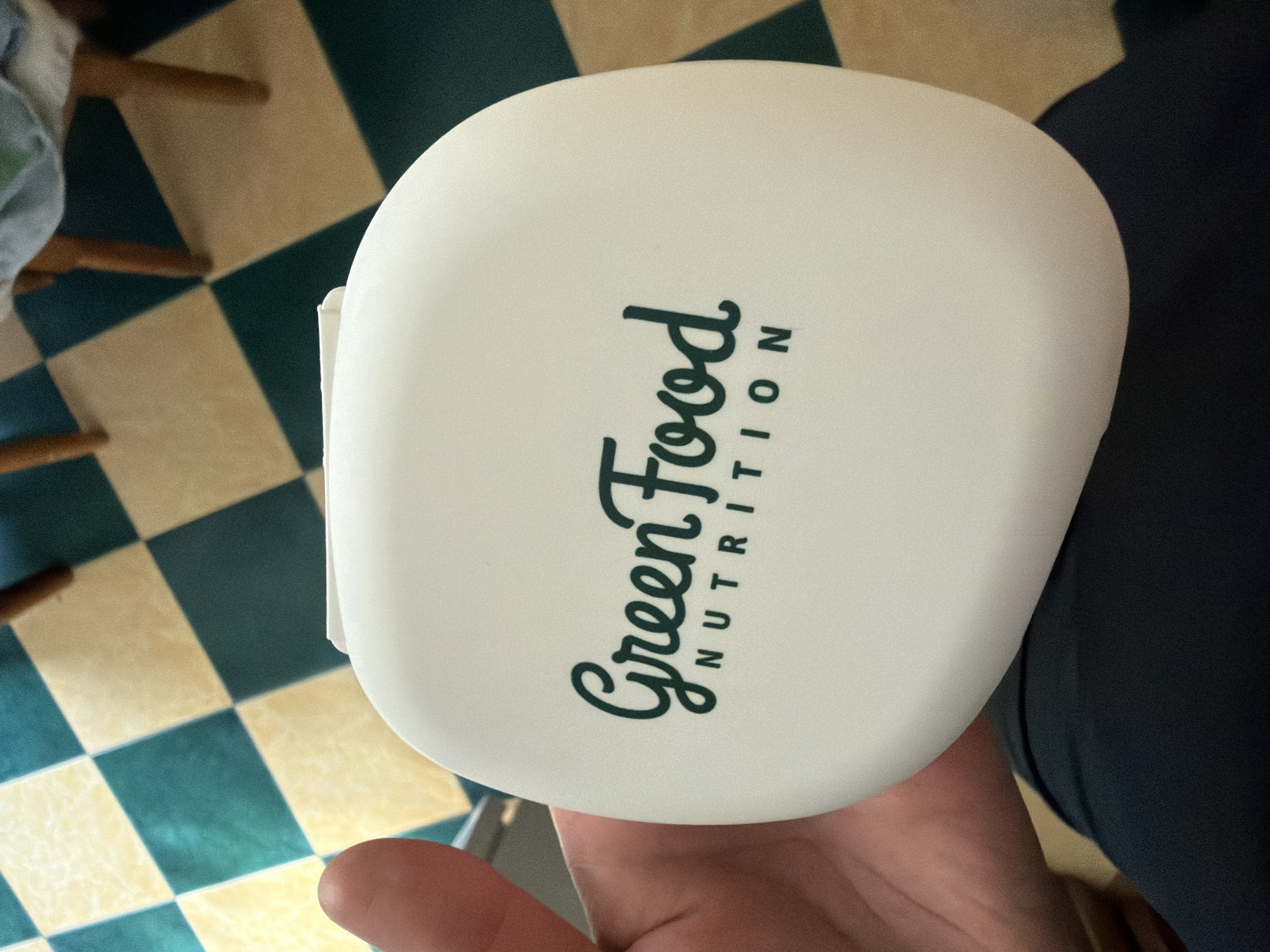 A close-up, eye-level shot shows a white, oval-shaped container with a dark green logo that reads "Green Food NUTRITION." The container is held by a person's hand, with their thumb visible on the left side of the frame. The container is resting on the person's dark blue pants. In the background, a checkered floor pattern of yellow and dark green tiles is visible, along with the legs of a wooden chair and a green and white cloth draped over it. The image appears to be taken indoors in an unknown location.