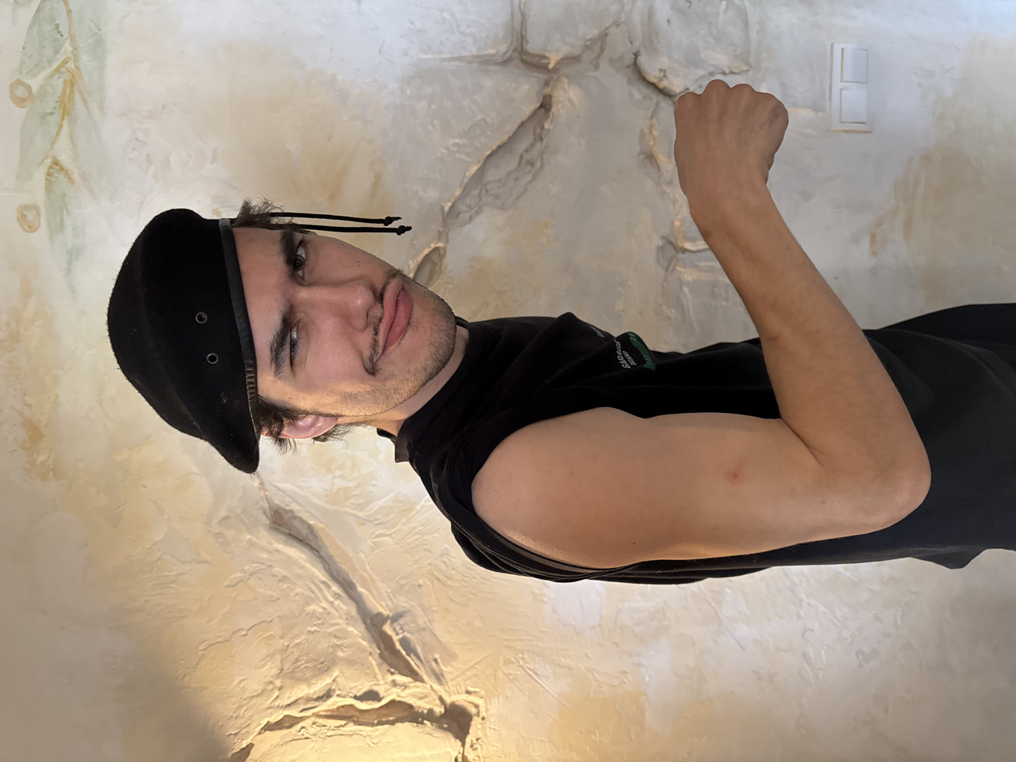 A man wearing a black beret and black sleeveless shirt is flexing his right arm in front of a light-colored wall with a textured surface. The man has short dark hair, a mustache, and a slight beard. He is looking at the camera with a somewhat exaggerated expression. A light switch is visible on the wall behind his arm. The location is not possible to identify from the image.