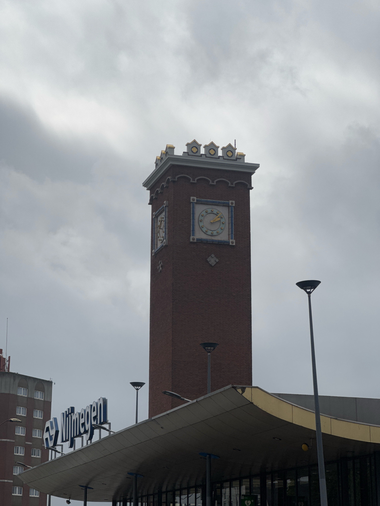 A brick clock tower with a decorative top stands tall against a cloudy sky. The clock face, featuring yellow hour markers and blue accents, shows the time to be approximately 9:40. Below the clock tower, a modern building with a curved, yellow-roofed entrance displays a large sign with the word "Nijmegen" in blue letters. To the left of the sign, another building with multiple floors and rows of windows is visible. Several lampposts, some with distinctive bowl-shaped tops, are scattered throughout the scene, and the overall atmosphere suggests an overcast day.