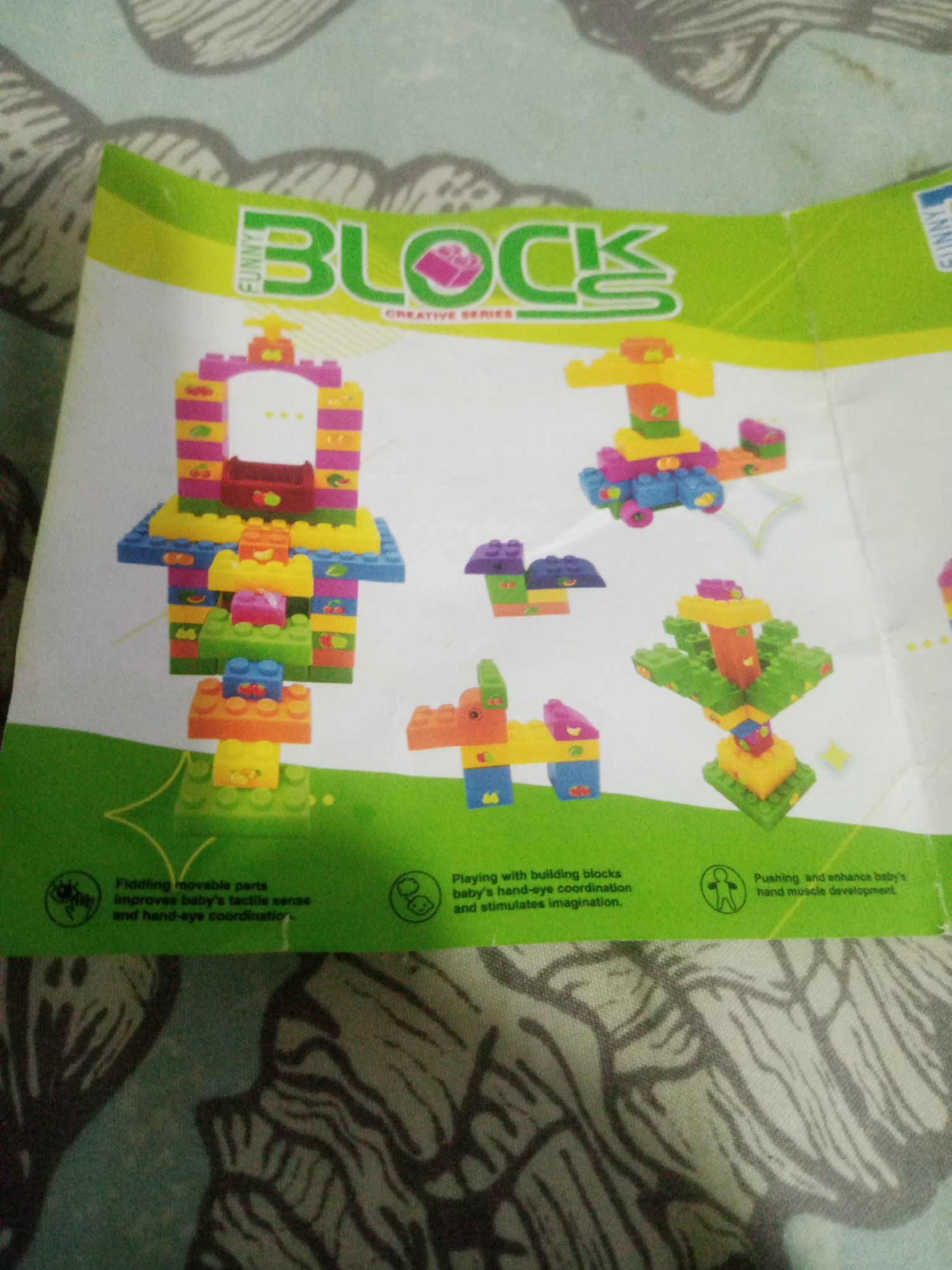 The image displays a colorful instruction booklet or packaging for a children's building block toy set, featuring various block structures. The booklet has a bright green border with text and icons. The main content showcases several assembled block models, including a castle-like structure, a vehicle, a tree, and animal figures. Below the toy models, there are text descriptions of the toy's benefits. The background of the image is a patterned fabric, possibly a tablecloth or blanket, with a light teal and gray design. The scene is indoors, and there is no indication of the specific location within La Tagua, Venezuela.