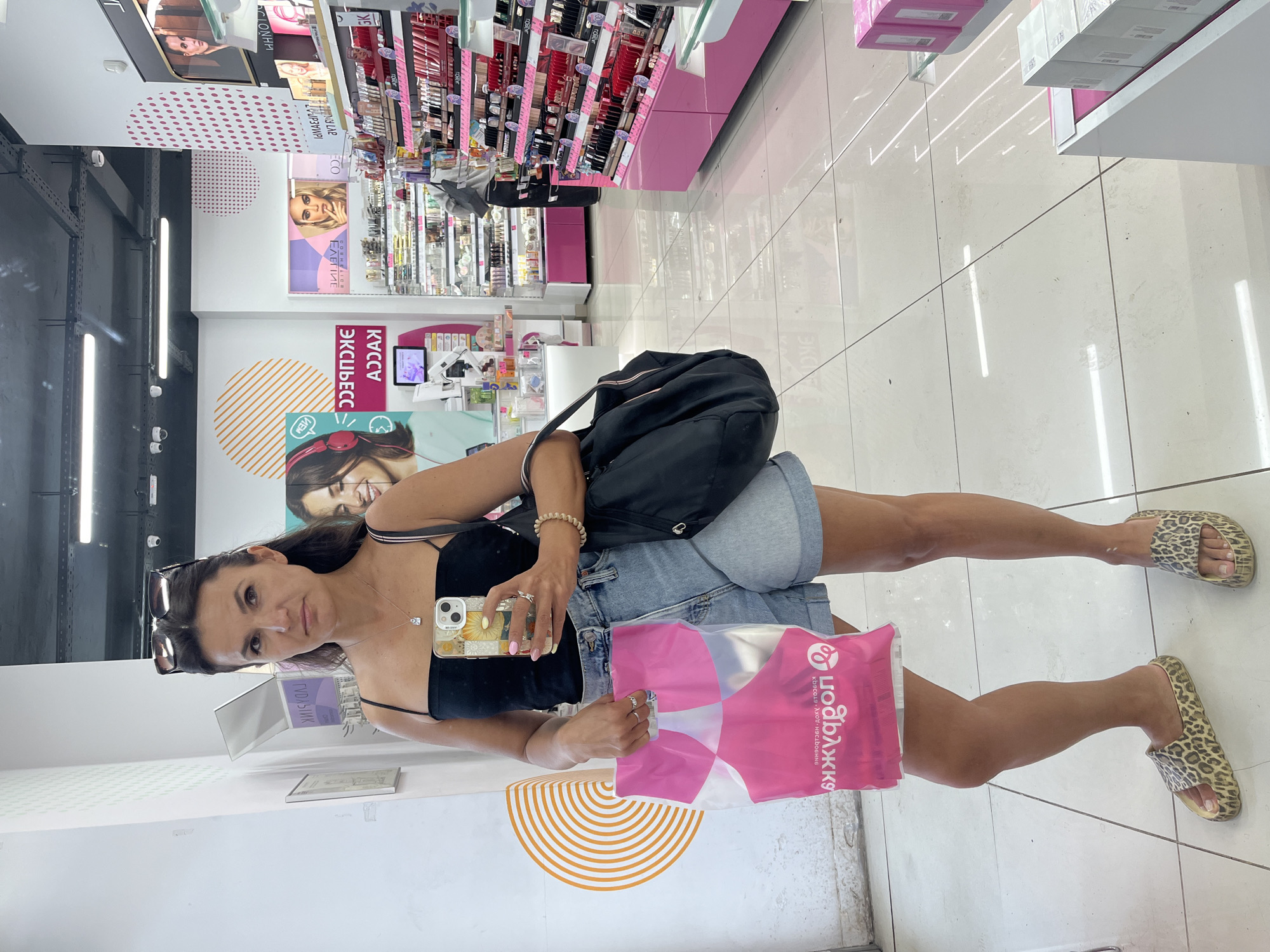 The image shows a woman standing in a cosmetics store, possibly in Moscow, Russia, as indicated by a logo on a shopping bag that appears to be "Rive Gauche," a well-known Russian cosmetics retailer. The woman is wearing a black tank top, denim shorts, and leopard-print slides. She is holding a pink shopping bag in her left hand and her smartphone in her right hand, which she is using to take a mirror selfie. She has sunglasses perched on top of her head and a delicate necklace with a pendant around her neck. The store's interior is visible, with shelves stocked with makeup and beauty products, bright lighting, and a tiled floor. There are also promotional posters and displays visible in the background.