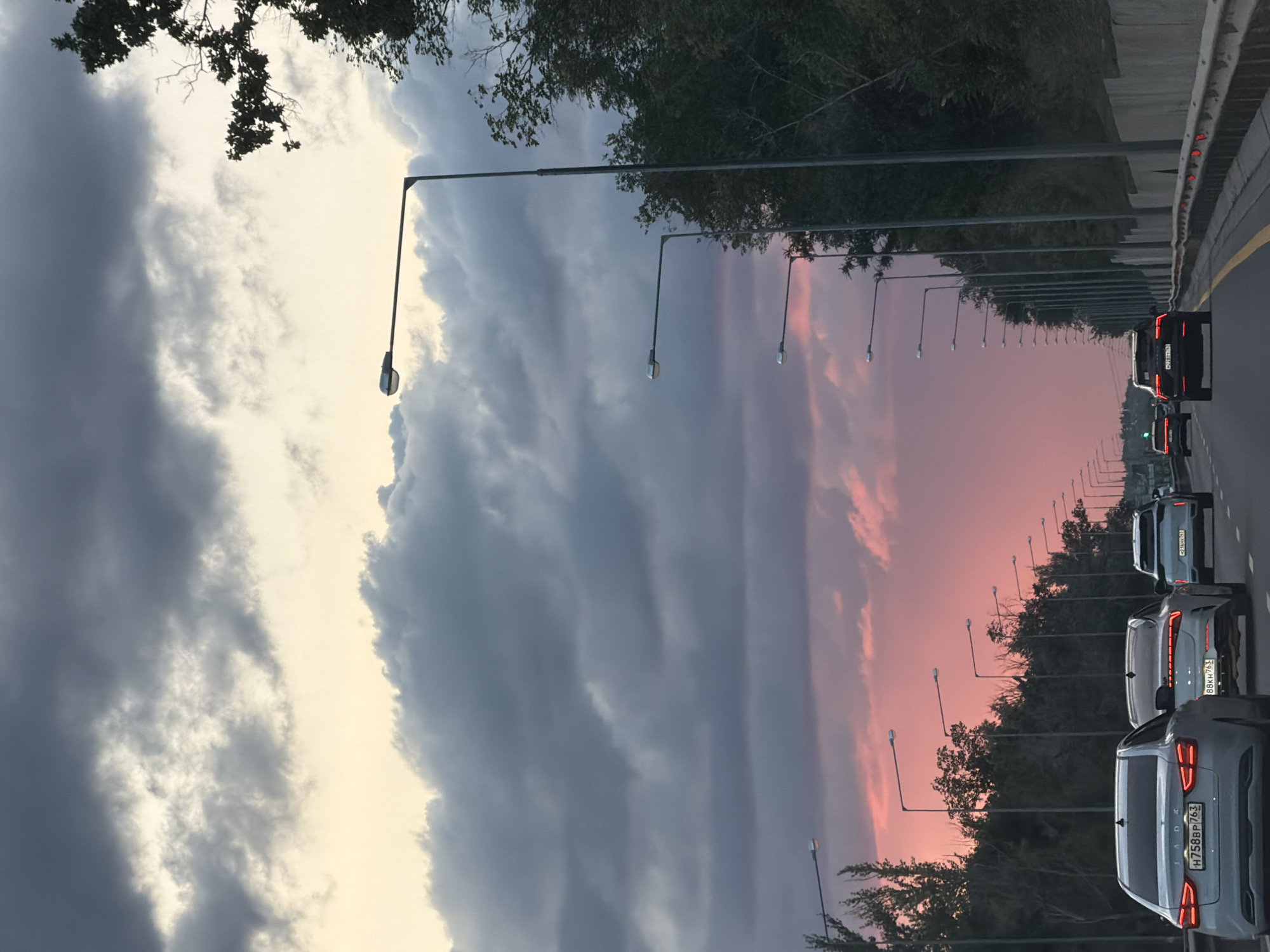 The image depicts a road in Samara, Russia, likely during sunset. A multi-lane highway is visible, with several cars traveling in the same direction. The sky is dramatic, with dark, heavy clouds occupying the upper portion of the frame, transitioning to a vibrant pink and orange hue closer to the horizon. Tall streetlights line the right side of the road, illuminated by the fading daylight. Dense trees and vegetation border the highway on the right, while the left side appears to have more sparse trees and foliage. The road surface is asphalt, with lane markings clearly visible. The overall impression is of a busy commute under an atmospheric sky.