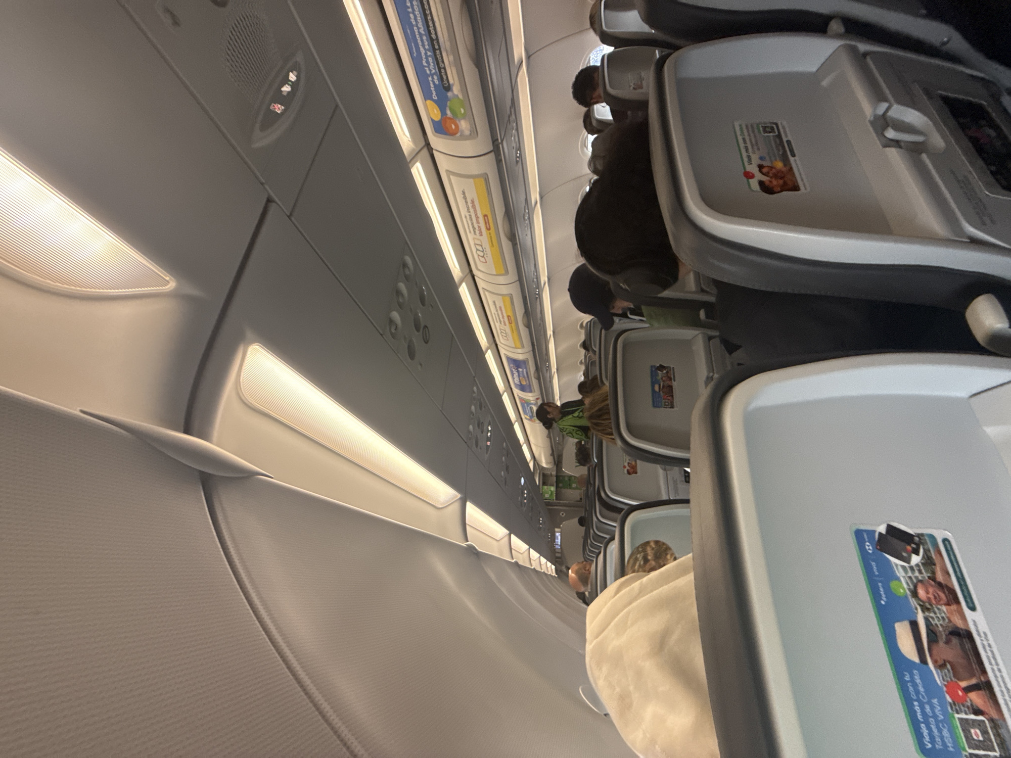 Here is a description of the image:
The image shows the interior of an airplane cabin, angled to emphasize the rows of seats and the overhead compartments. Passengers are seated in their respective chairs. Overhead, lights illuminate the ceiling panels, which display various signs and advertisements. The seatbacks feature entertainment screens and promotional stickers.