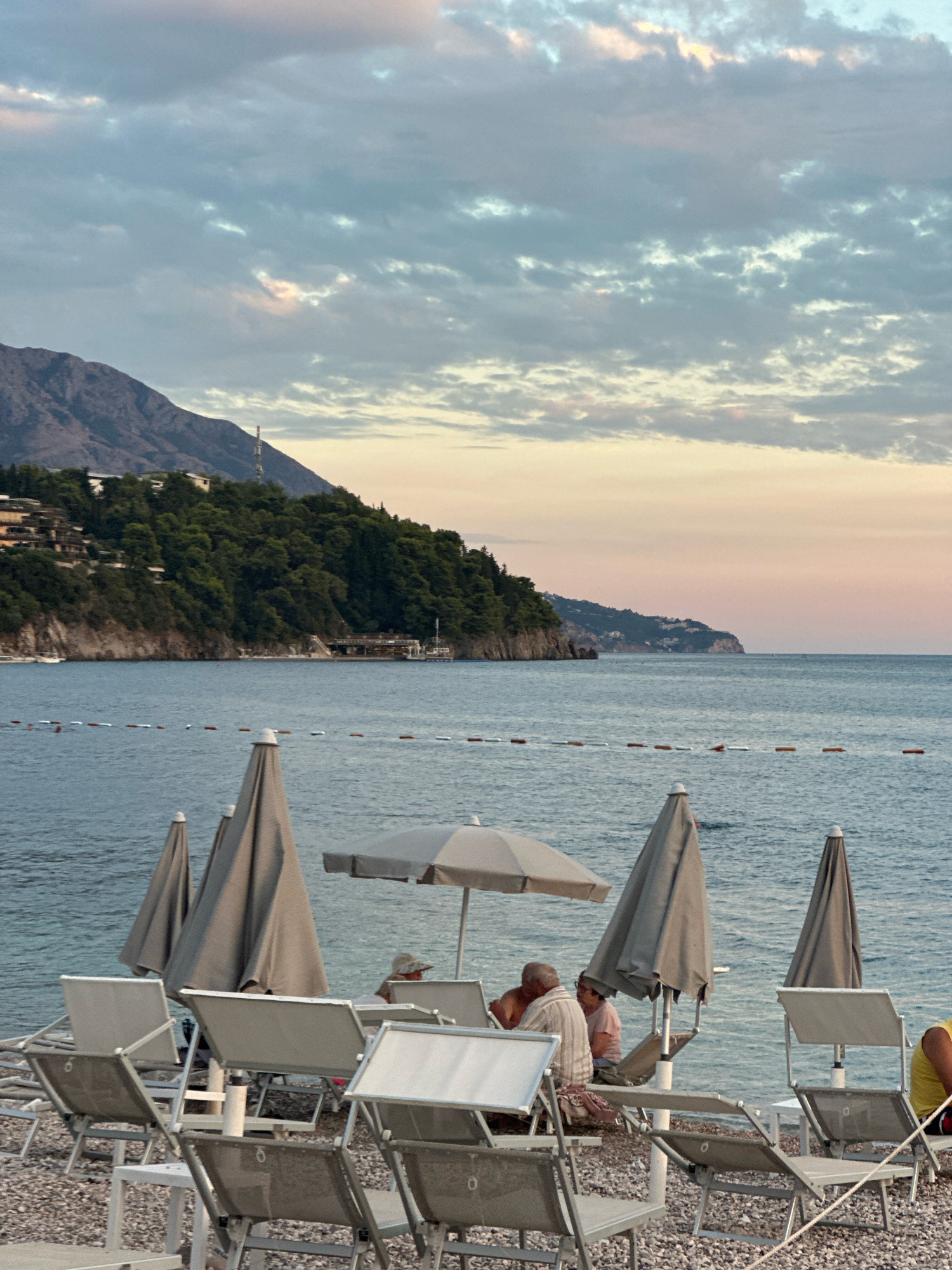 The image depicts a serene beach scene at dusk in Budva, Montenegro. In the foreground, numerous empty beach chairs and closed umbrellas are arranged on a pebbled shore. Several people are seated under a large umbrella, appearing to be engaged in conversation or enjoying the view.
The middle ground features the calm blue sea, marked by a line of buoys stretching across the water, indicating a swimming area. Beyond the sea, a lush green hillside rises, dotted with buildings and trees, characteristic of Budva's coastal landscape. The hill slopes down to the water, with some rocky outcrops visible.
The background is dominated by a vast sky filled with soft, wispy clouds that are beginning to take on the warm hues of sunset, transitioning from pale pink to a soft orange and blue. A distant mountain range can be seen on the left, adding to the dramatic backdrop. The overall atmosphere is peaceful and relaxing, capturing the end of a day at the beach.